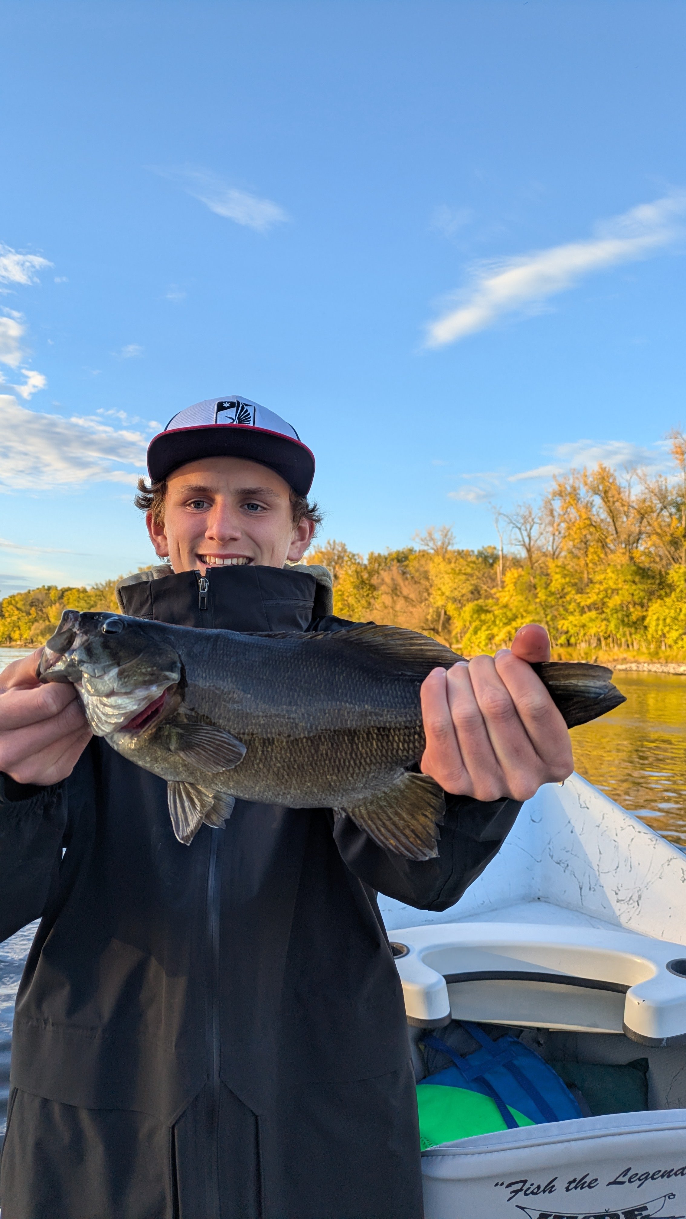 A young man in a black jacket and a baseball cap holding a large fish on a boat with water, trees, and a blue sky in the background.