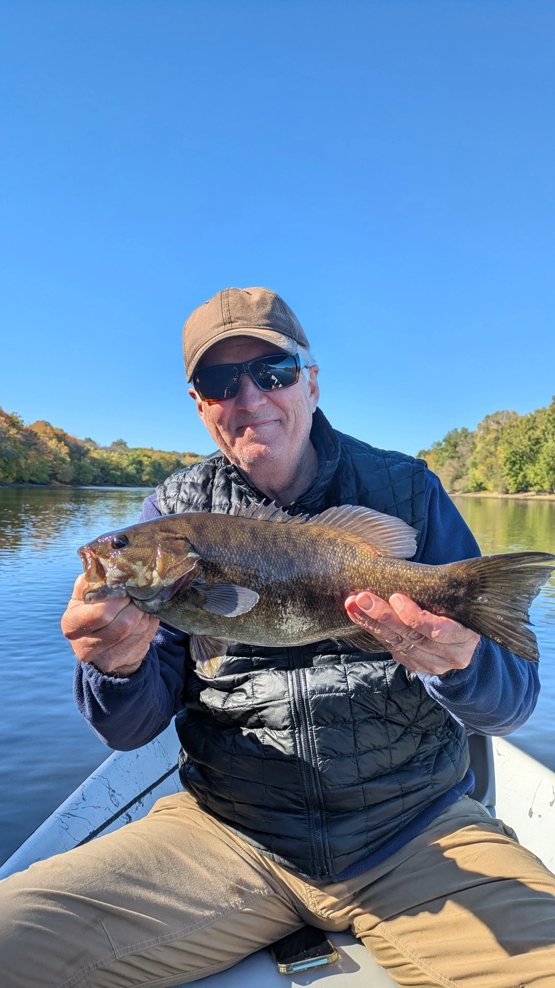 A man wearing sunglasses, a cap, a black quilted vest, and khaki pants, sitting in a boat on a river, holding a large fish with both hands, with trees and a clear blue sky in the background.