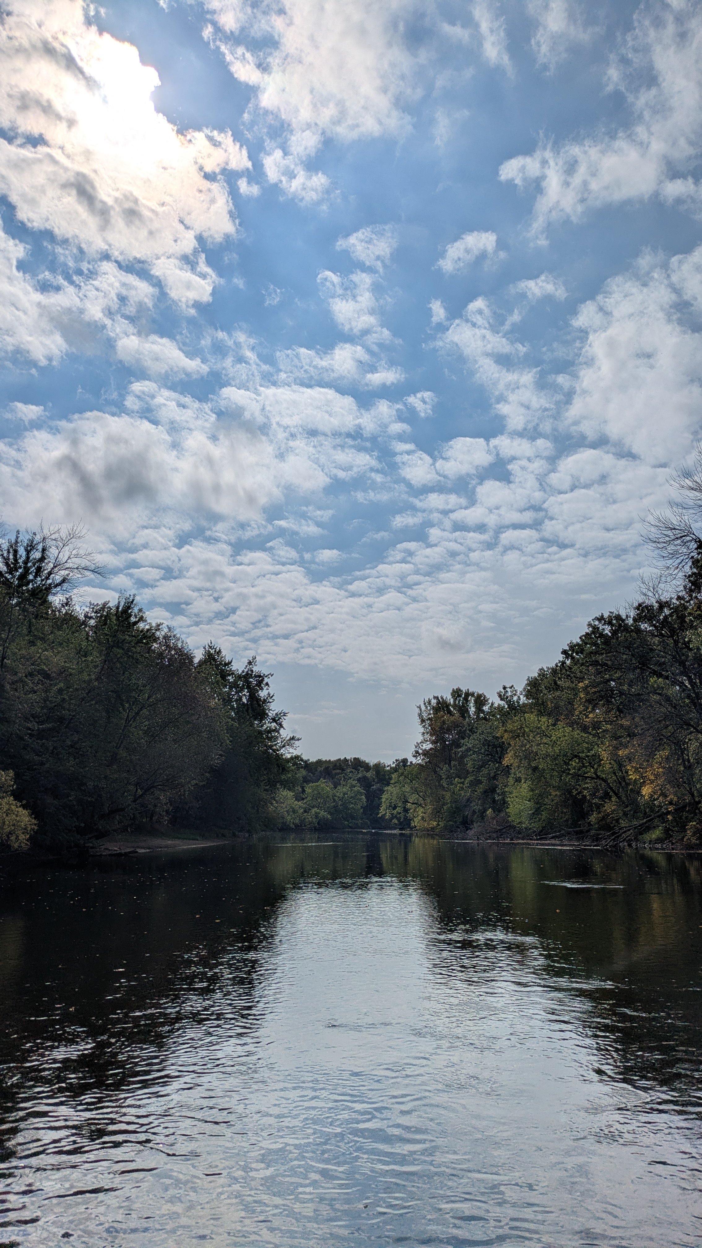A calm river flowing through a wooded area under a partly cloudy sky.