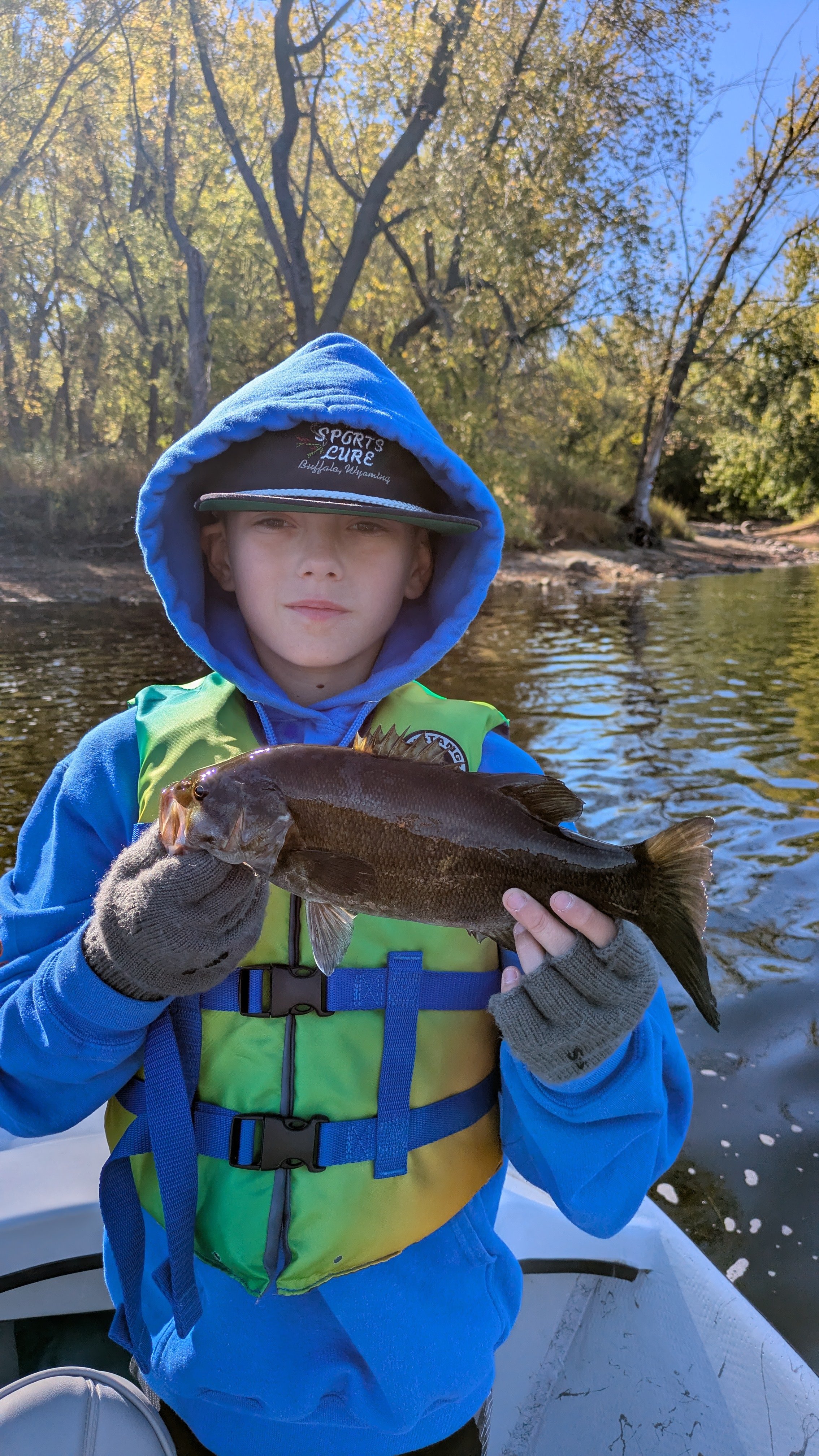 A young boy wearing a blue hoodie and light green life jacket holding a fish while standing on a boat near the water, with trees and the river in the background.