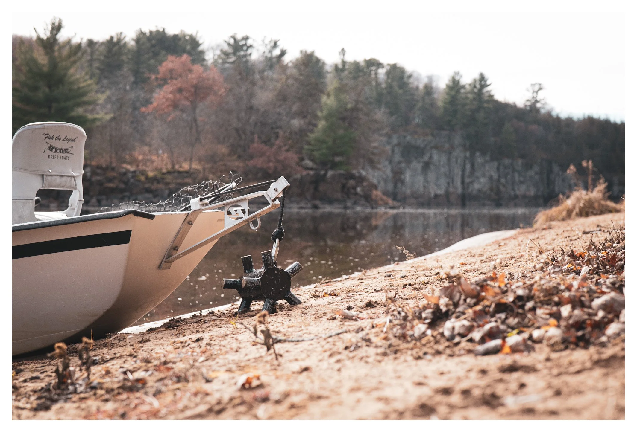A boat with a white seat and a black and white hull is beached on sandy shore near a body of water. There is a black cleat on the sand and a steel cable on the boat. Trees with autumn foliage are in the background.