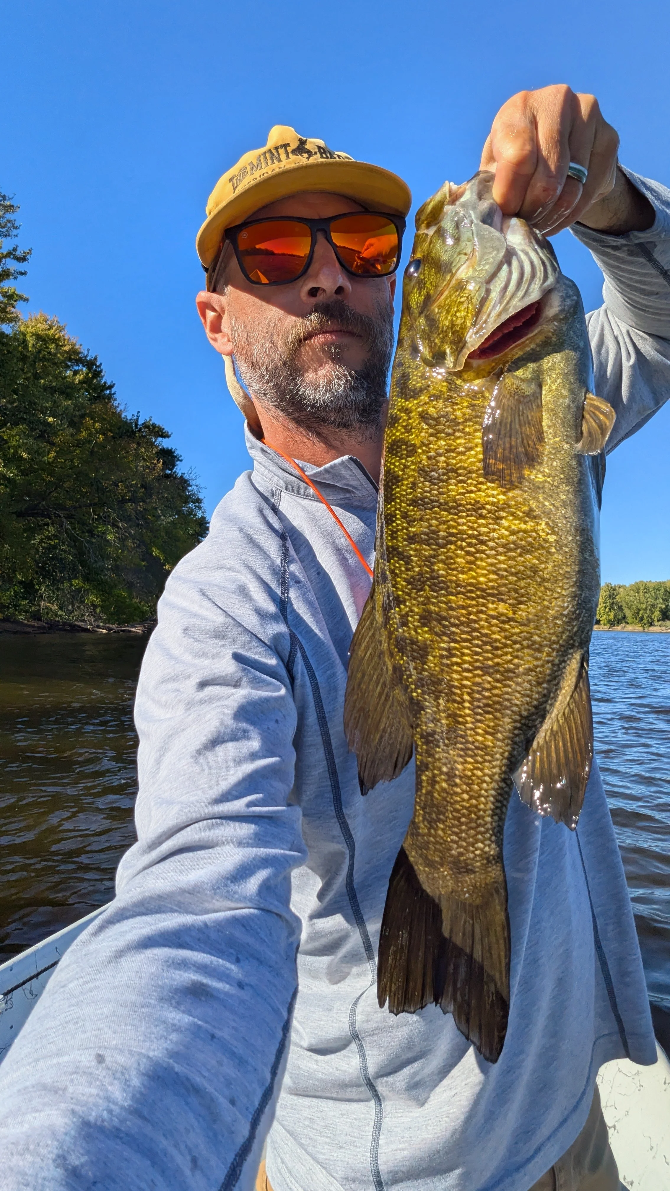 A man wearing sunglasses and a yellow cap holding a large fish over a body of water, with trees and blue sky in the background.