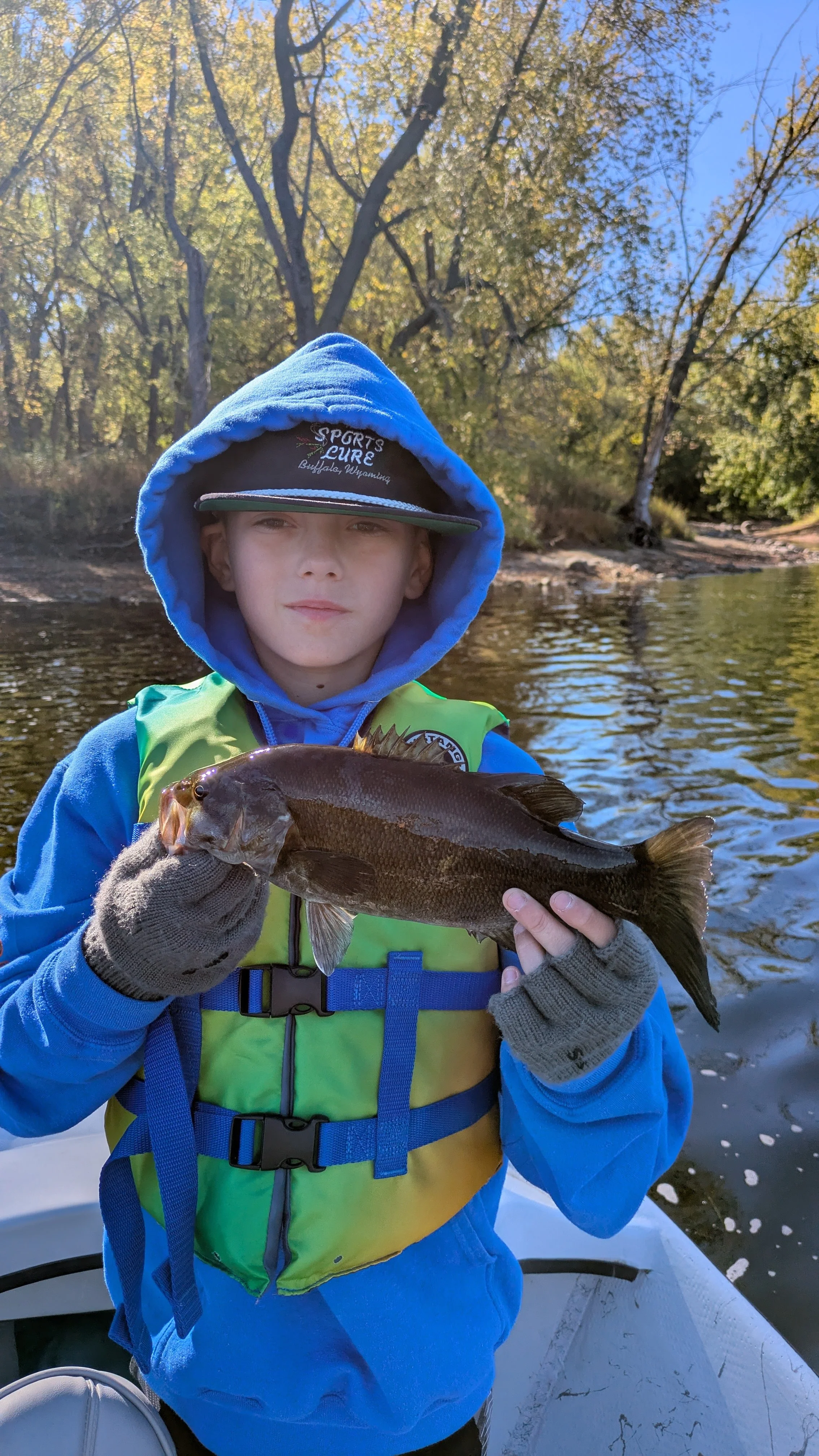 A boy wearing a blue hoodie, a green life vest, and gloves, holding a fish he caught while on a boat in a river, with trees and a clear sky in the background.
