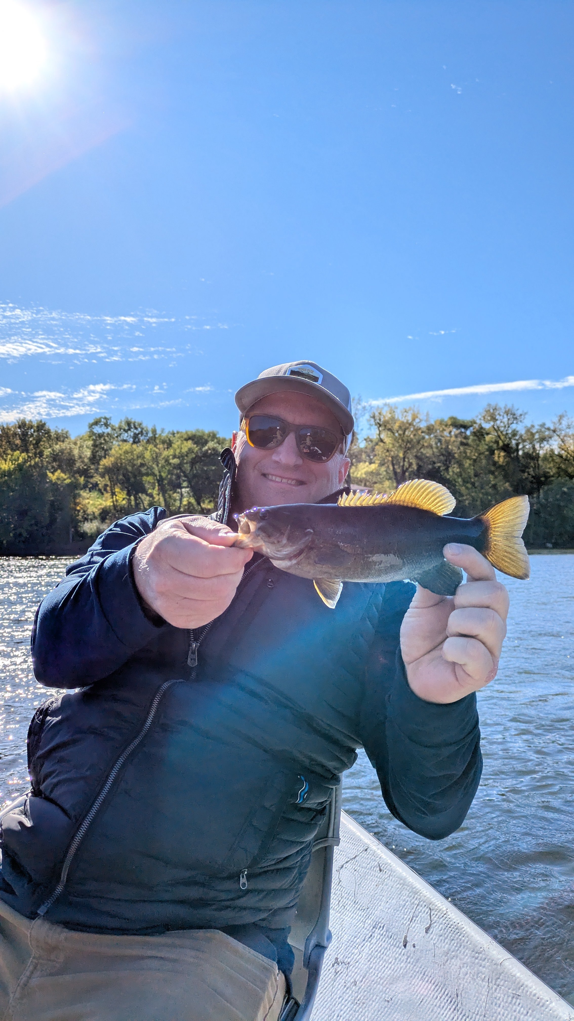 A man smiling and wearing sunglasses and a cap, holding a fish he caught while on a boat on a lake with trees in the background, under a blue sky.