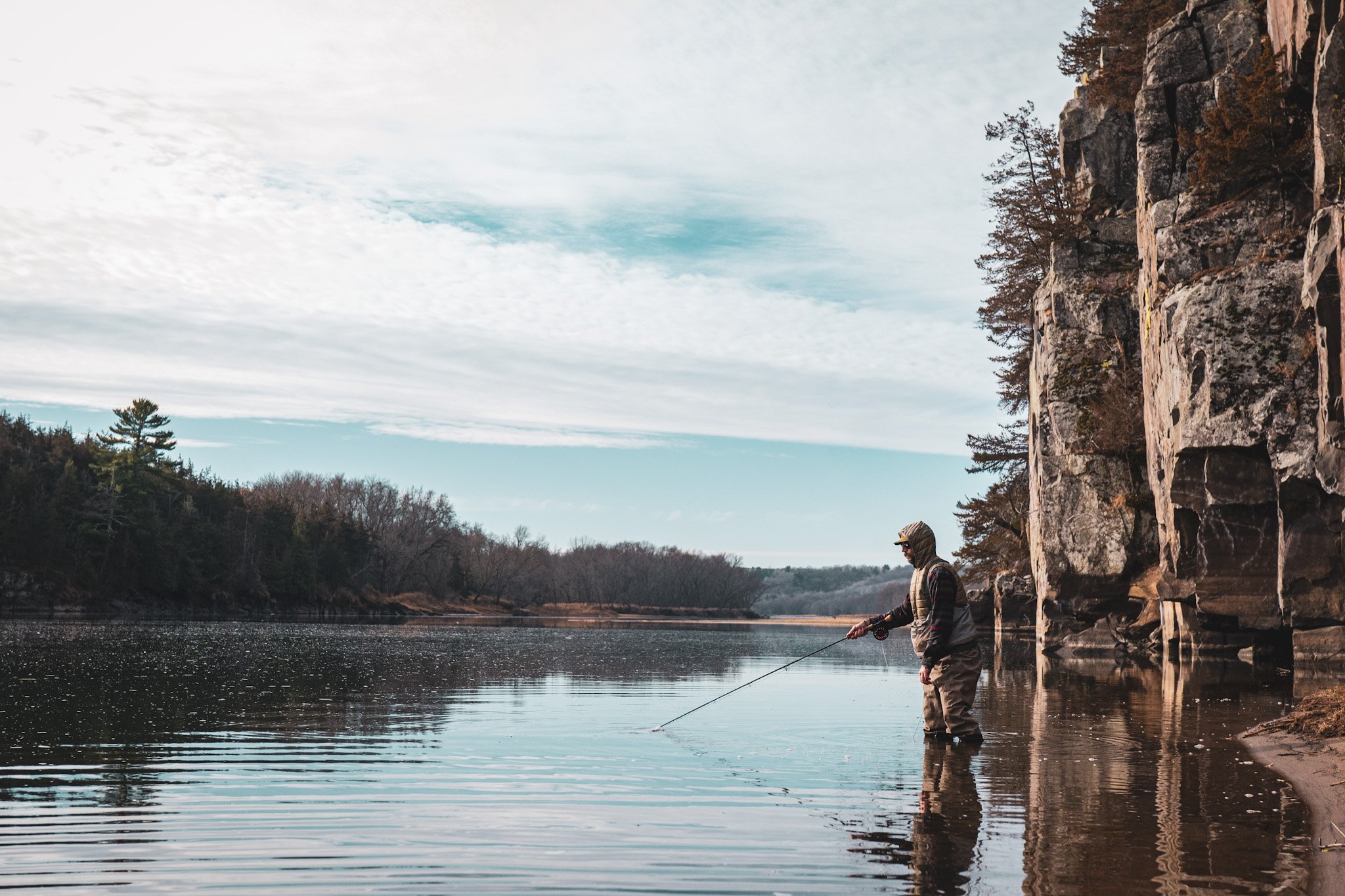 A person fishing in a river near rocky cliffs and trees under a cloudy sky.