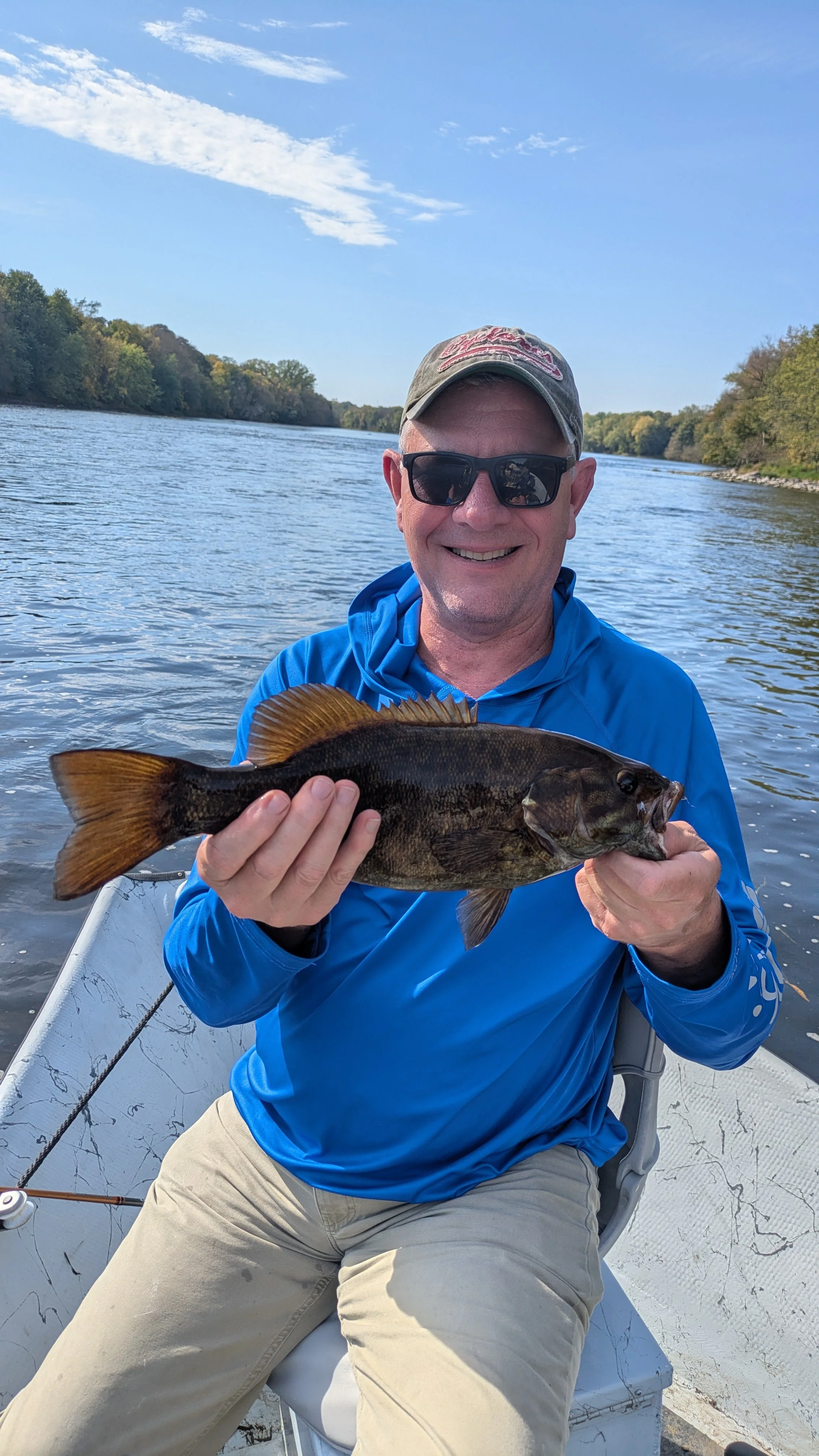 A man wearing sunglasses and a blue jacket sitting in a boat on a river, holding a fish he caught, with trees and a partly cloudy sky in the background.