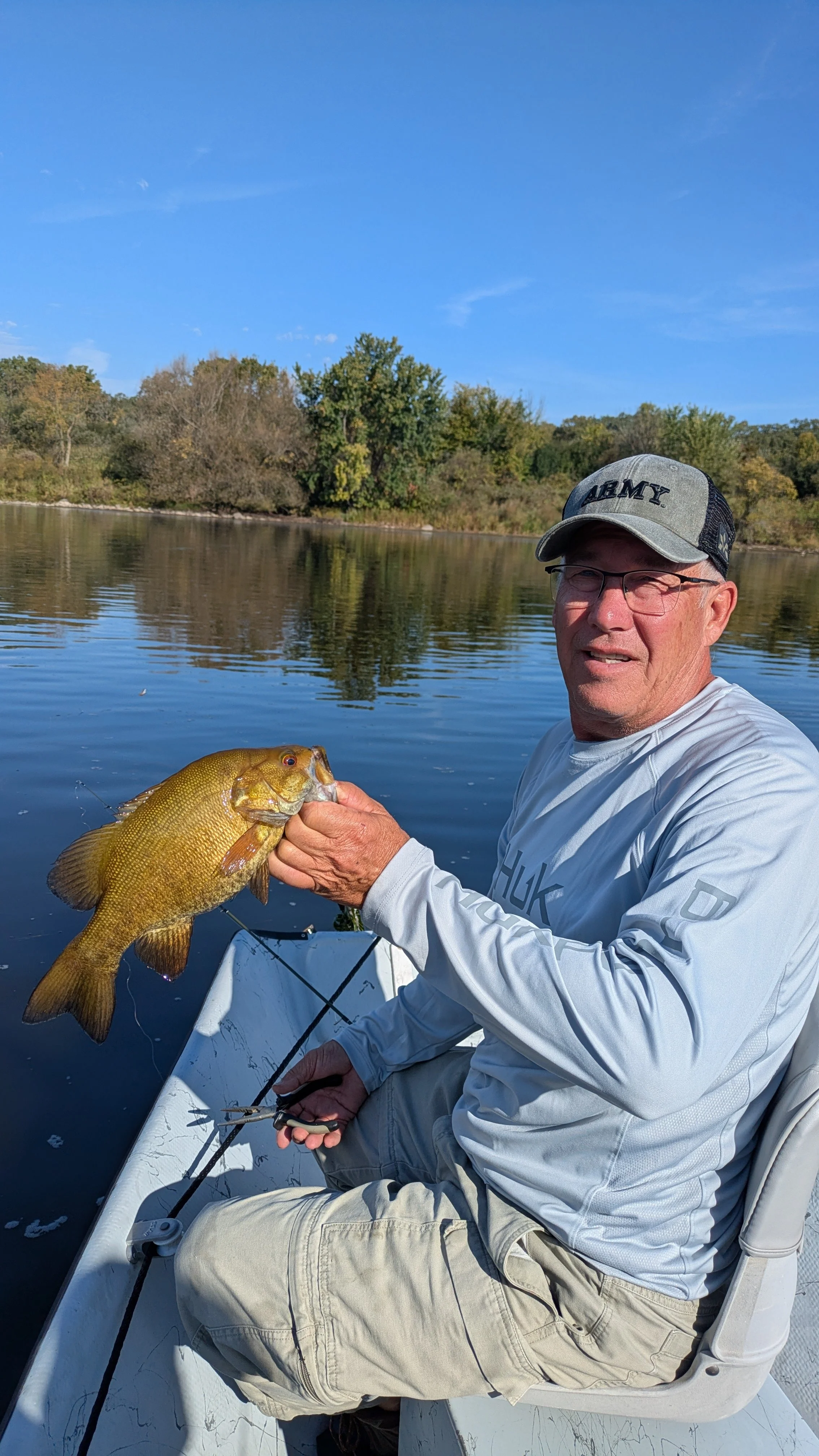 A man wearing a gray cap and glasses sitting on a boat, holding a fish he caught with a fishing rod by the water, with trees and blue sky in the background.
