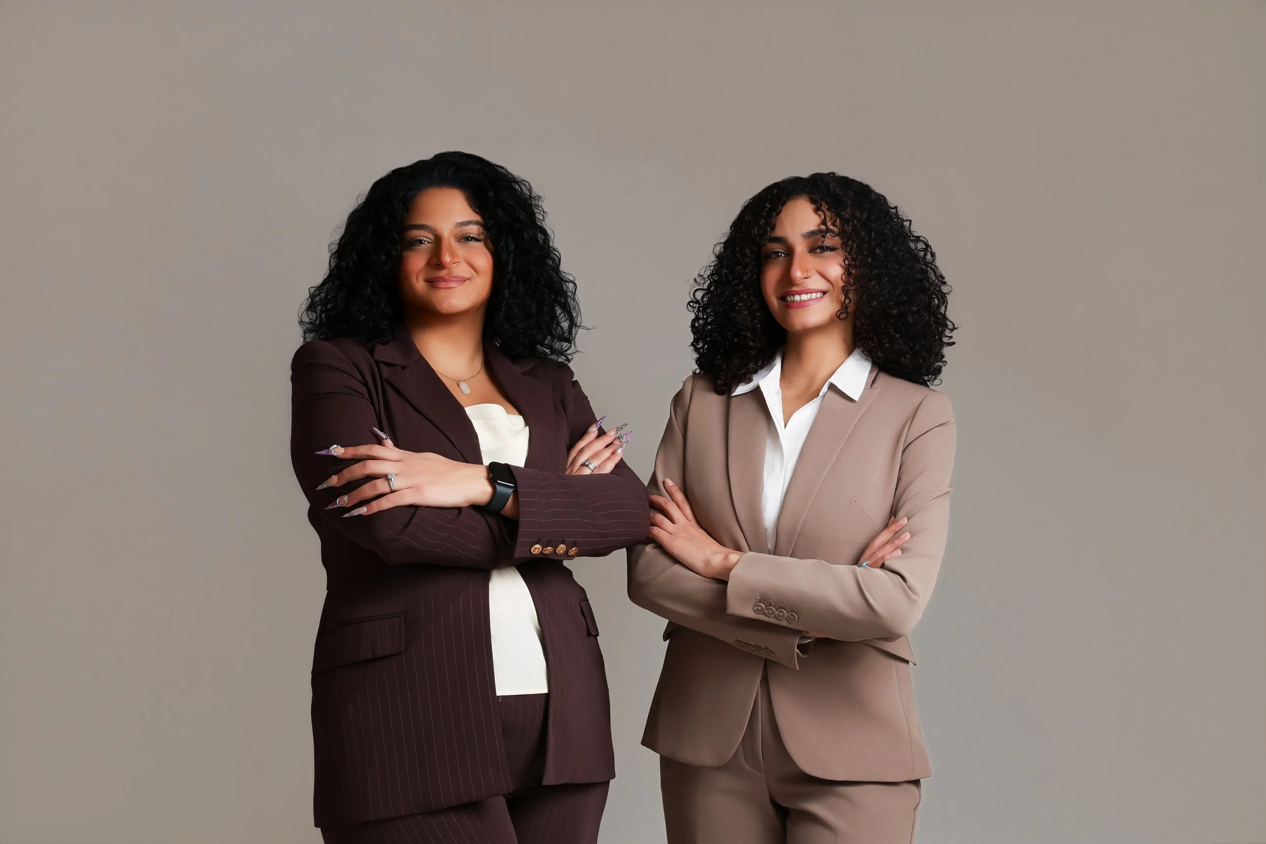 Two professional women standing with arms crossed, smiling, dressed in business suits, against a plain gray background.