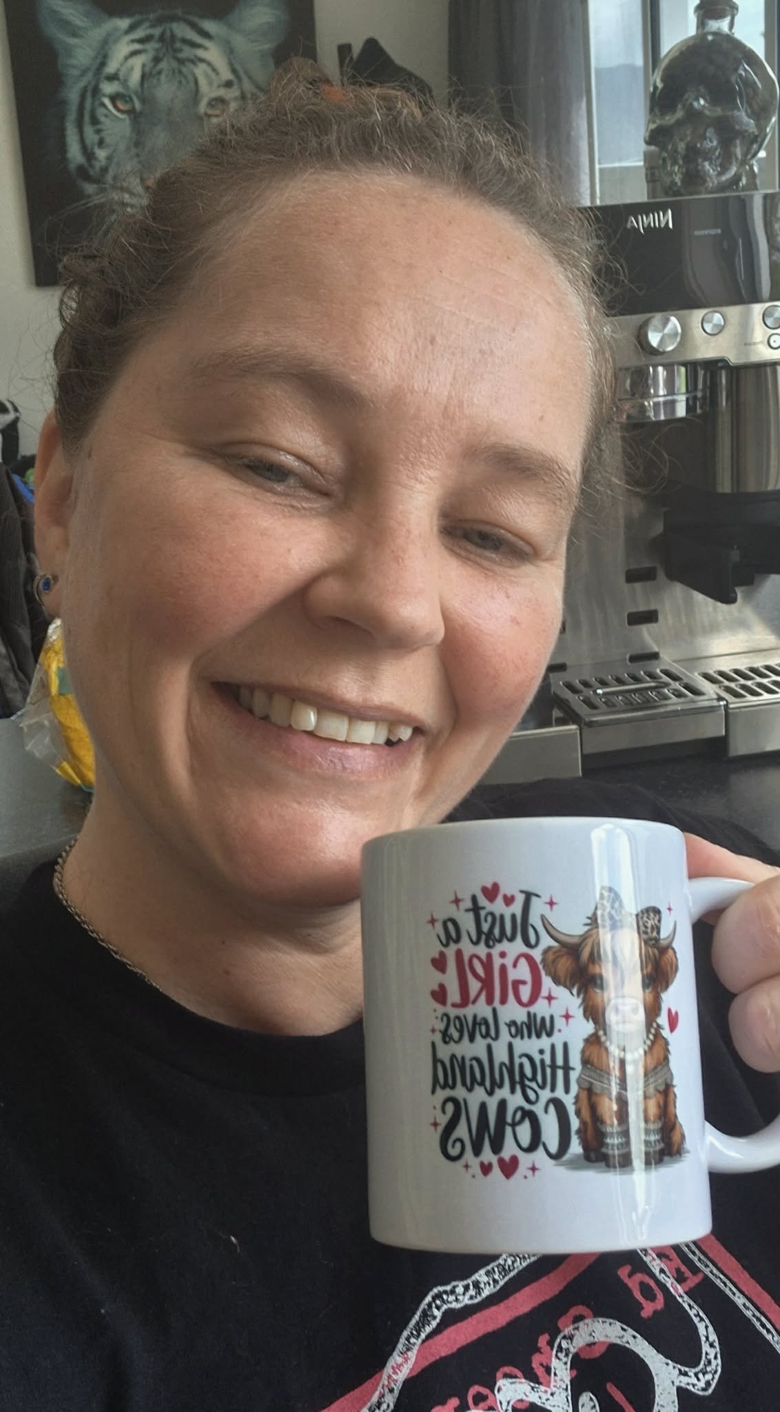 owner of Coffee and Moo holding a mug that has a colorful design and text that reads 'That Girl who loves her Highland Cows' with an illustration of a Highland Cow on it. In the background, there is a coffee machine and a tiger painting on the wall.