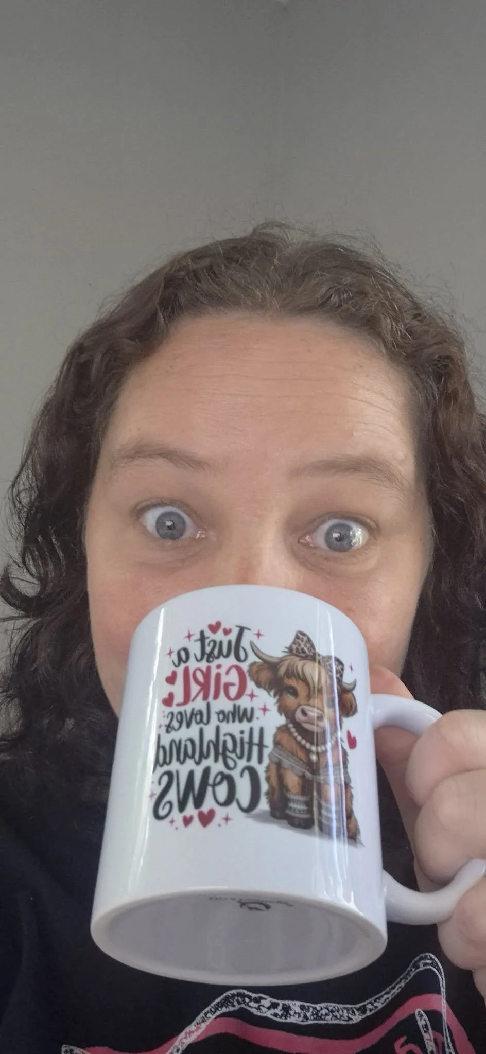 Owner of Coffee and Moo holding a white mug with a cow illustration and text saying "Just a girl who loves hilarious cows" in front of her face, partially obscuring her mouth. She has wide-open eyes and is looking at the camera.