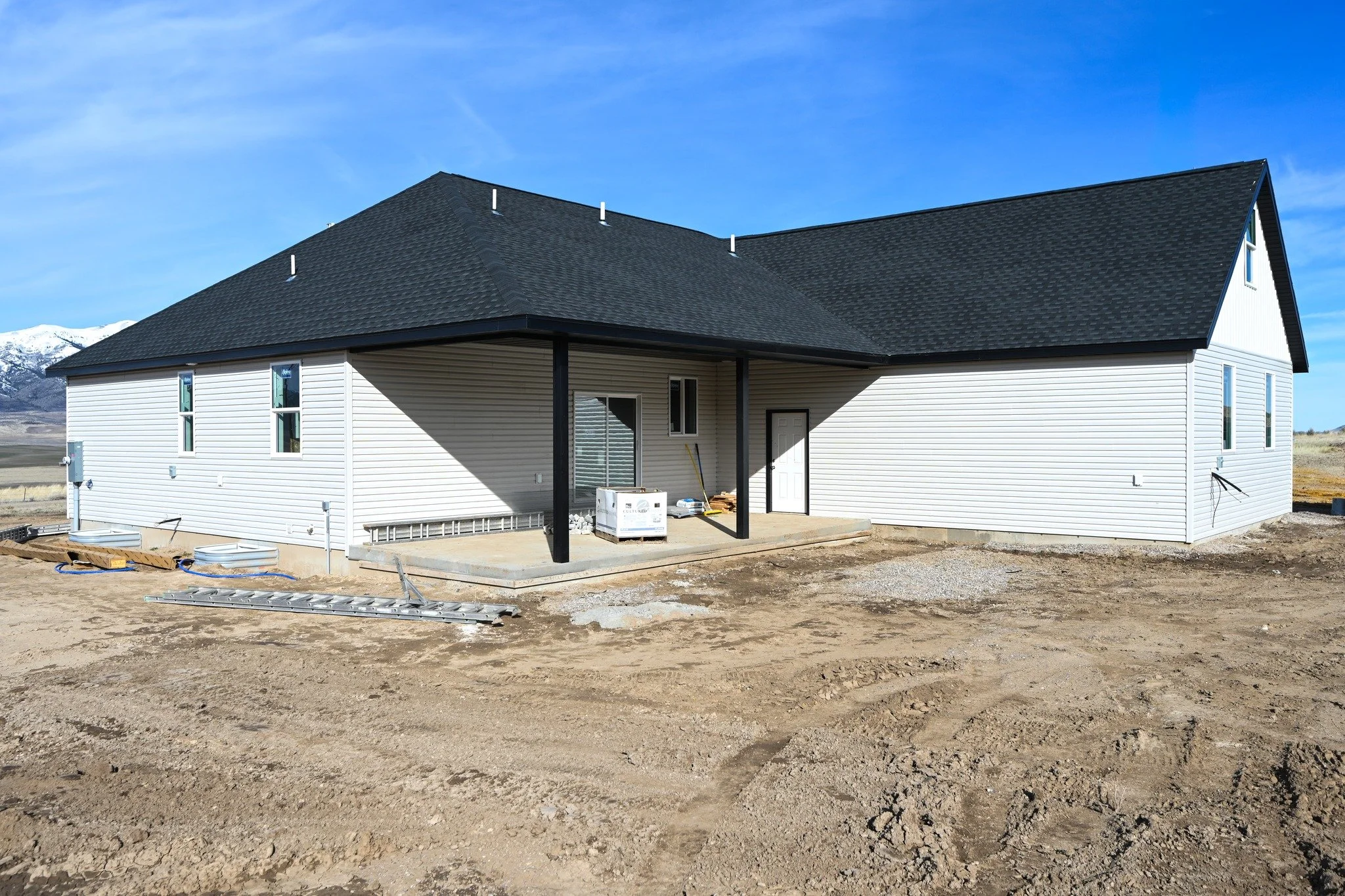 Newly built house with white siding and black roof on a dirt lot, with construction materials and tools on the porch. Background shows mountains and a blue sky.