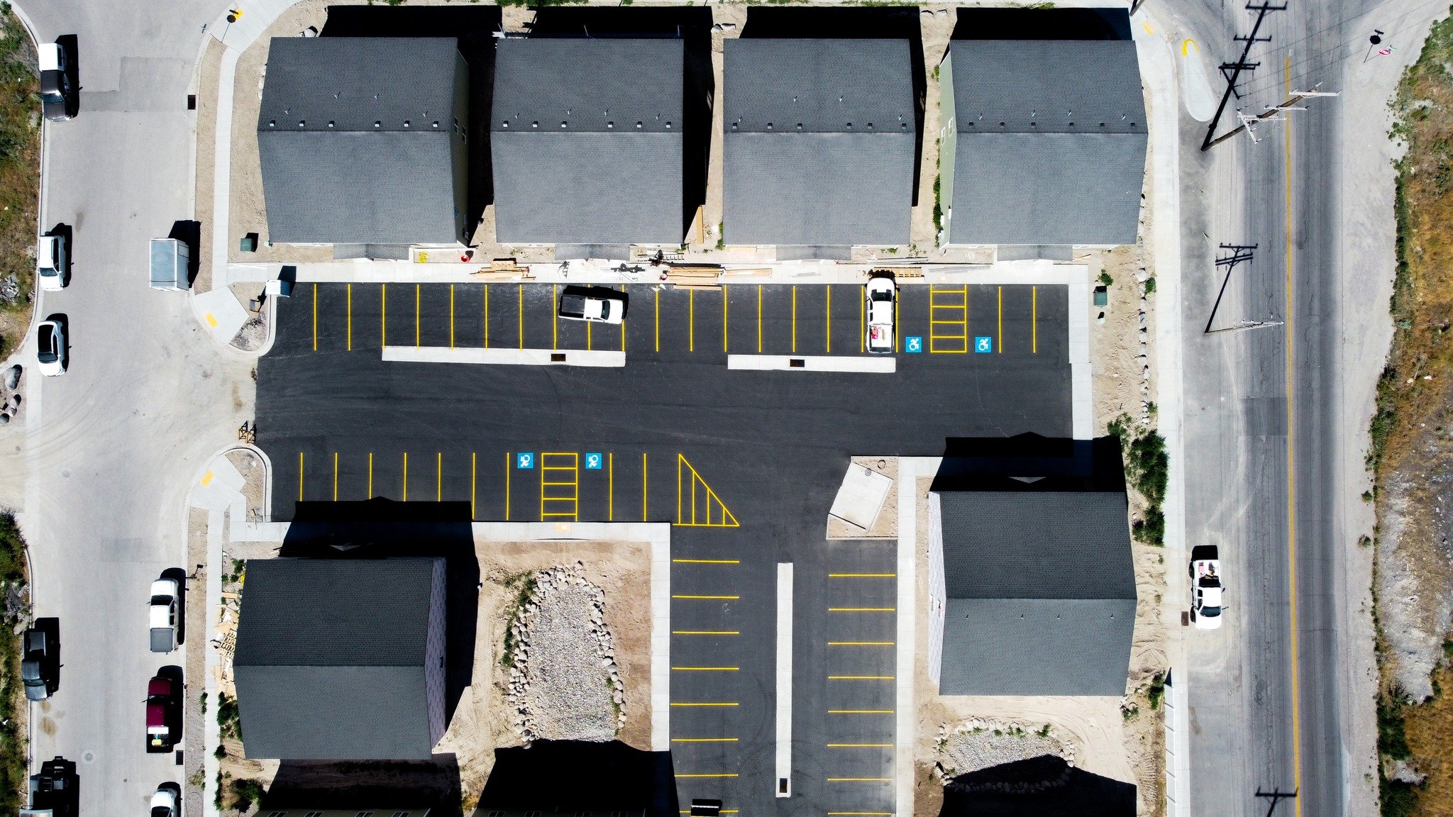 A newly paved parking lot with marked handicap parking spaces, surrounded by several buildings, with a few cars parked and some construction or landscaping in progress.