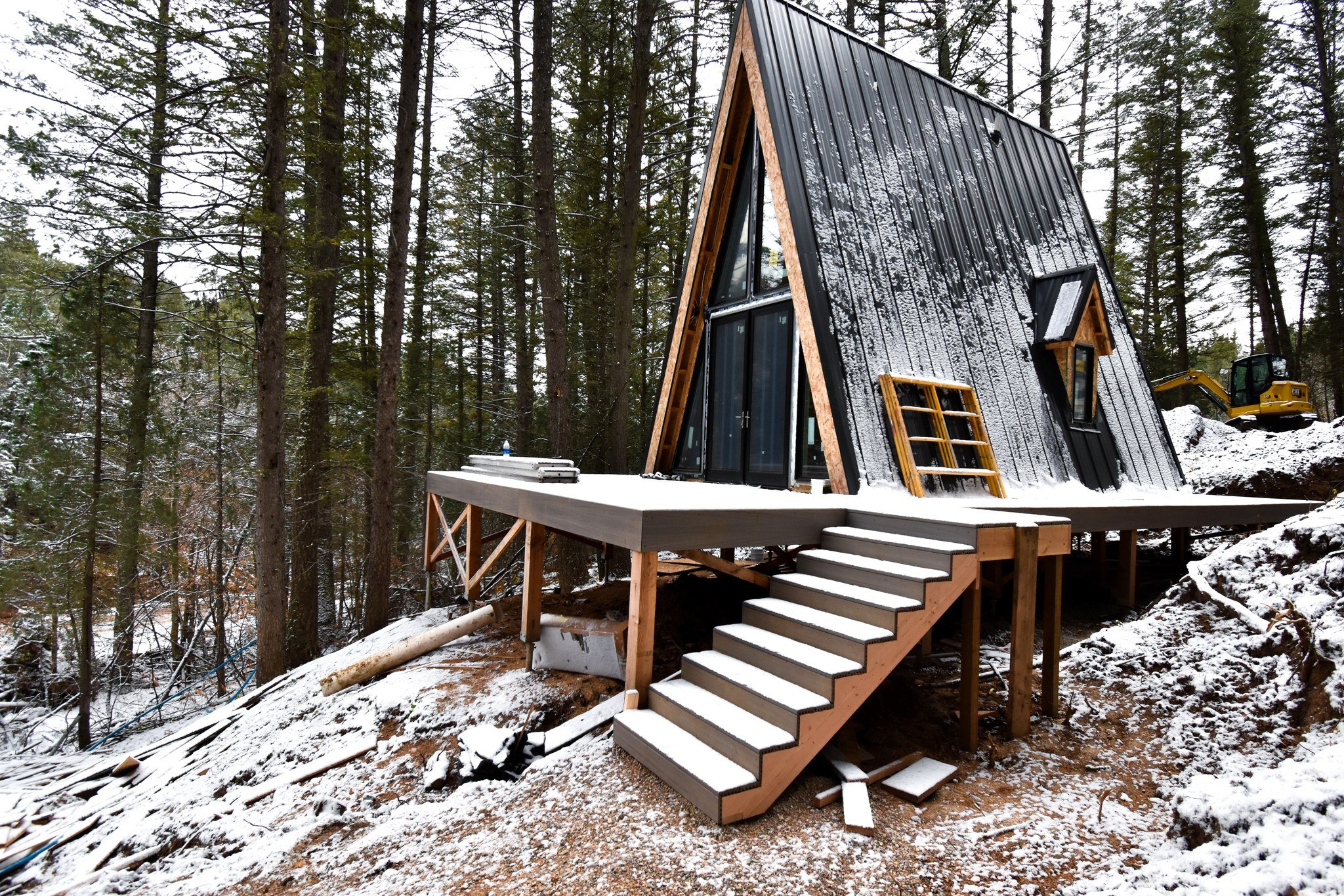 A black A-frame cabin with a snowy roof is elevated on a wooden deck with stairs in a forested area during winter.