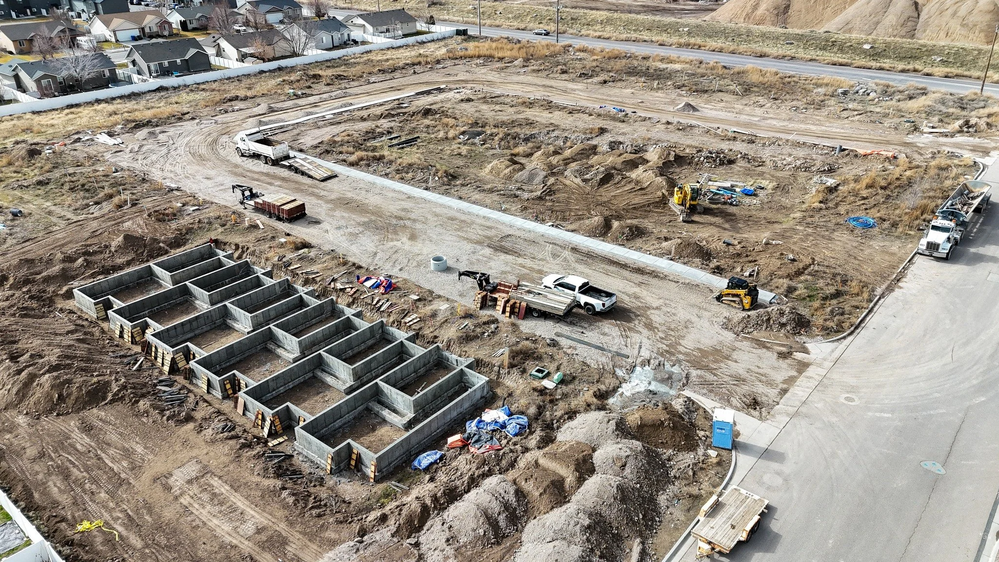Construction site with concrete foundation walls and building materials, surrounded by dirt and gravel, with construction vehicles and equipment present.