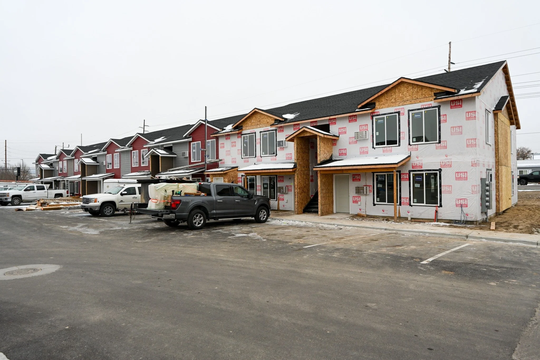 Row of multi-story houses under construction with some covered in siding and others with exposed insulation, parked trucks and construction materials in the foreground, overcast sky.