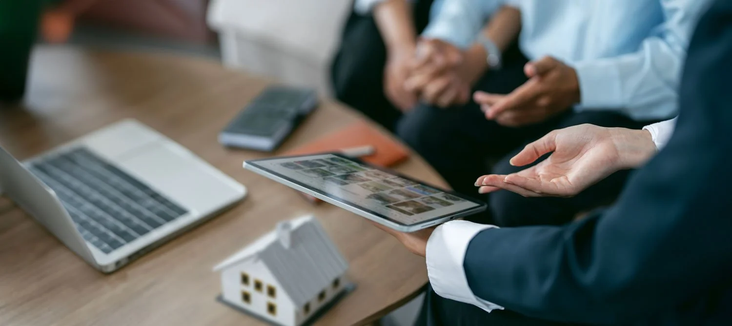 Person in business attire holding a tablet with pictures, sitting at a desk with a laptop, remote control, notebooks, and a small house model during a meeting.