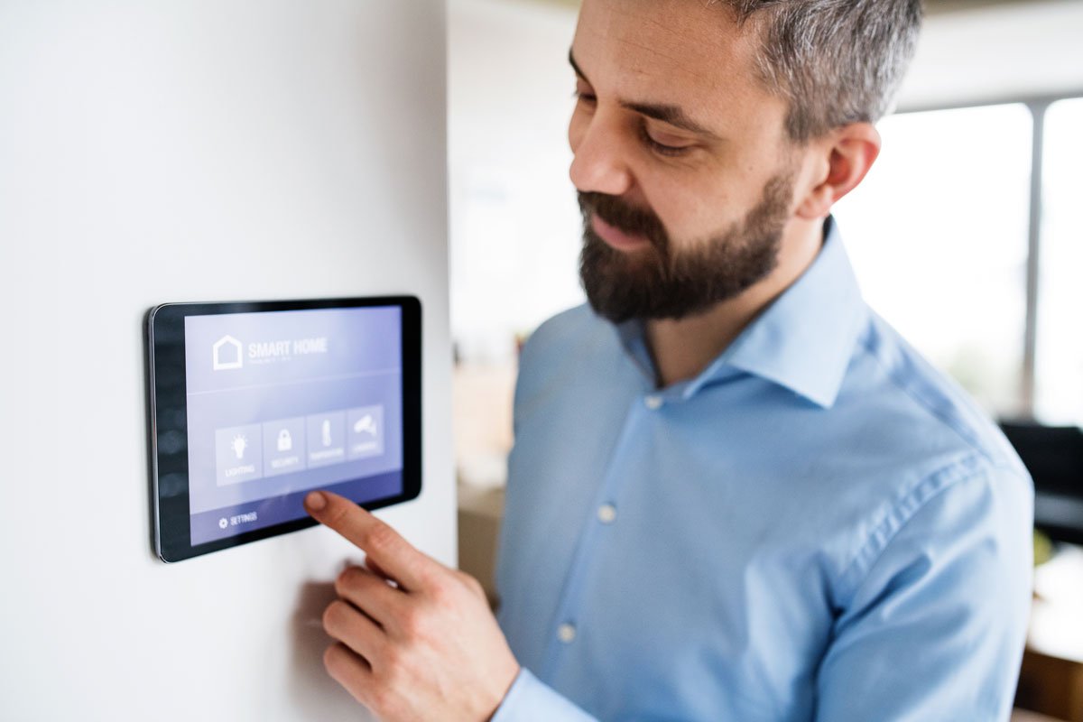 A man in a light blue shirt interacts with a wall-mounted smart home control panel in a bright room.