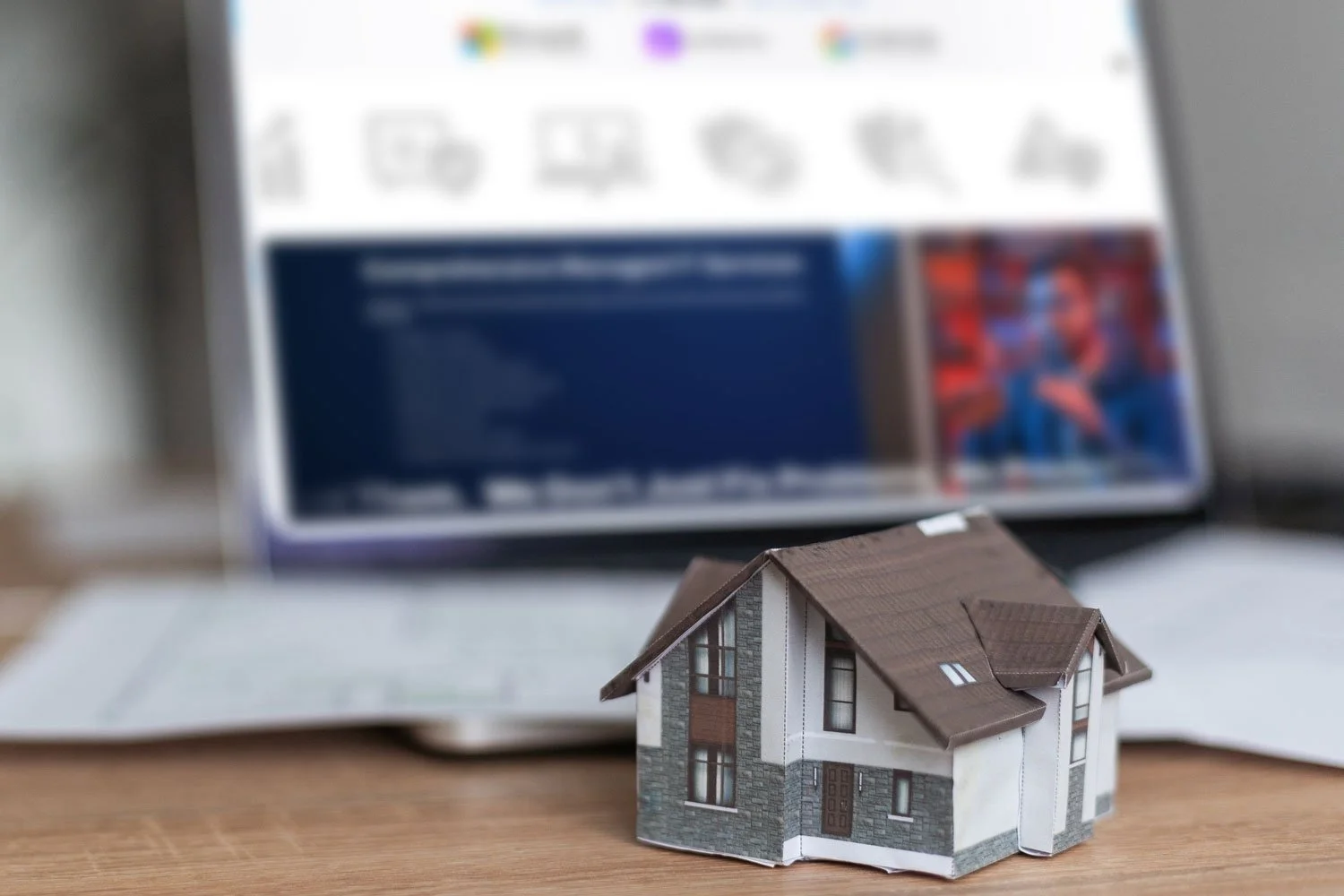 A small, paper model of a house on a desk with a blurred tablet in the background displaying a website.
