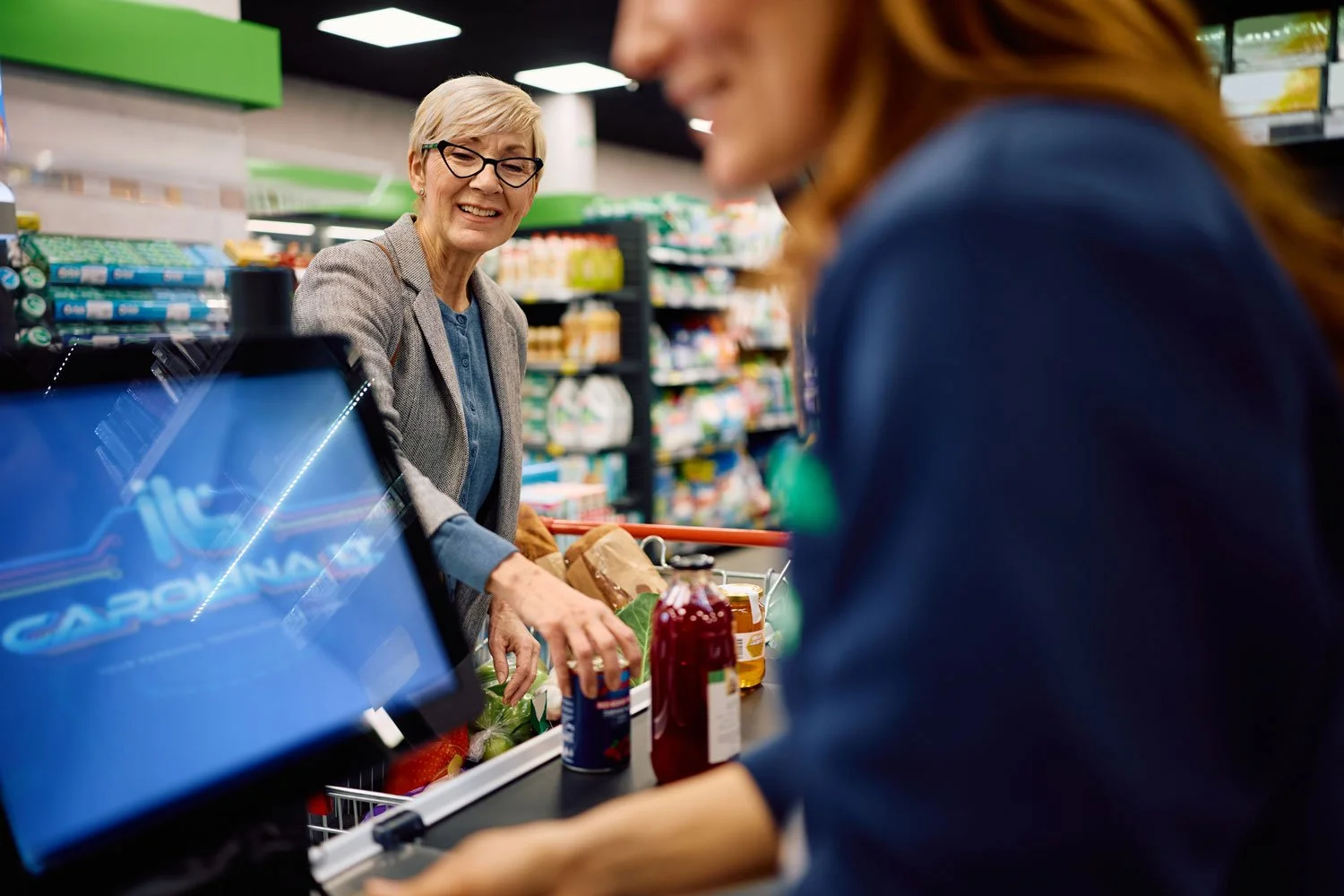 A woman with glasses and short blond hair smiling at a cashier, who is a red-haired woman in a blue jacket, in a grocery store checkout line. The checkout counter has bottles of ketchup and mustard, and groceries in a paper bag.