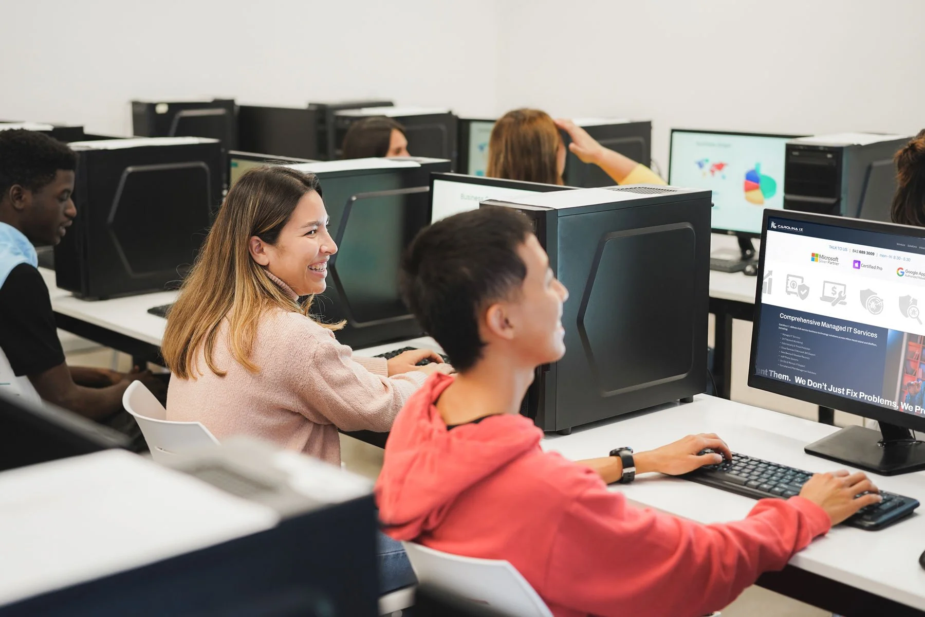 A diverse group of students in a computer lab, smiling and working on desktop computers, with some screens displaying colorful graphs and a website about IT services.