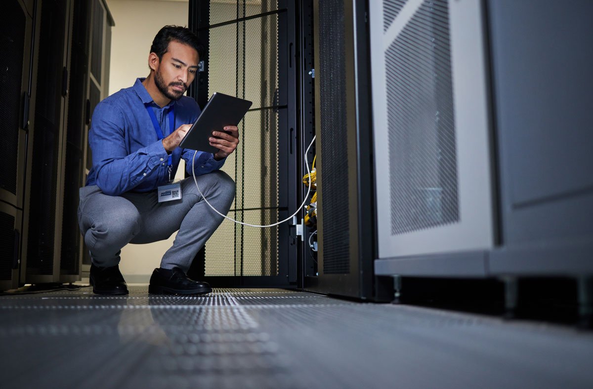 A technician crouching in a server room checking a tablet near server racks.