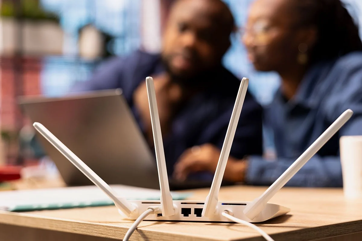 Wireless router with four antennas on a table, with two people blurred in the background looking at a tablet.