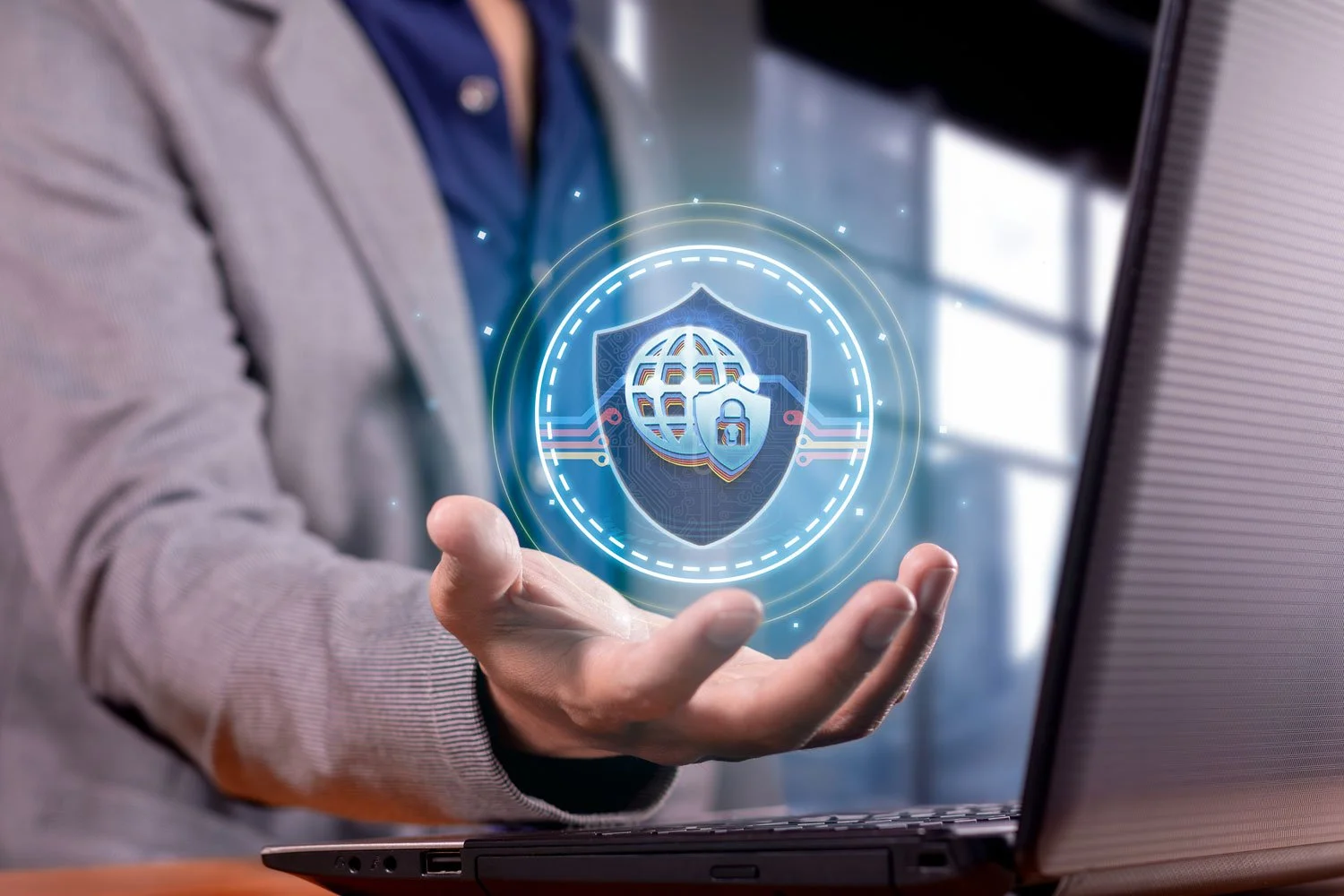 Person holding a holographic digital security icon with a shield, globe, and padlock over a laptop.