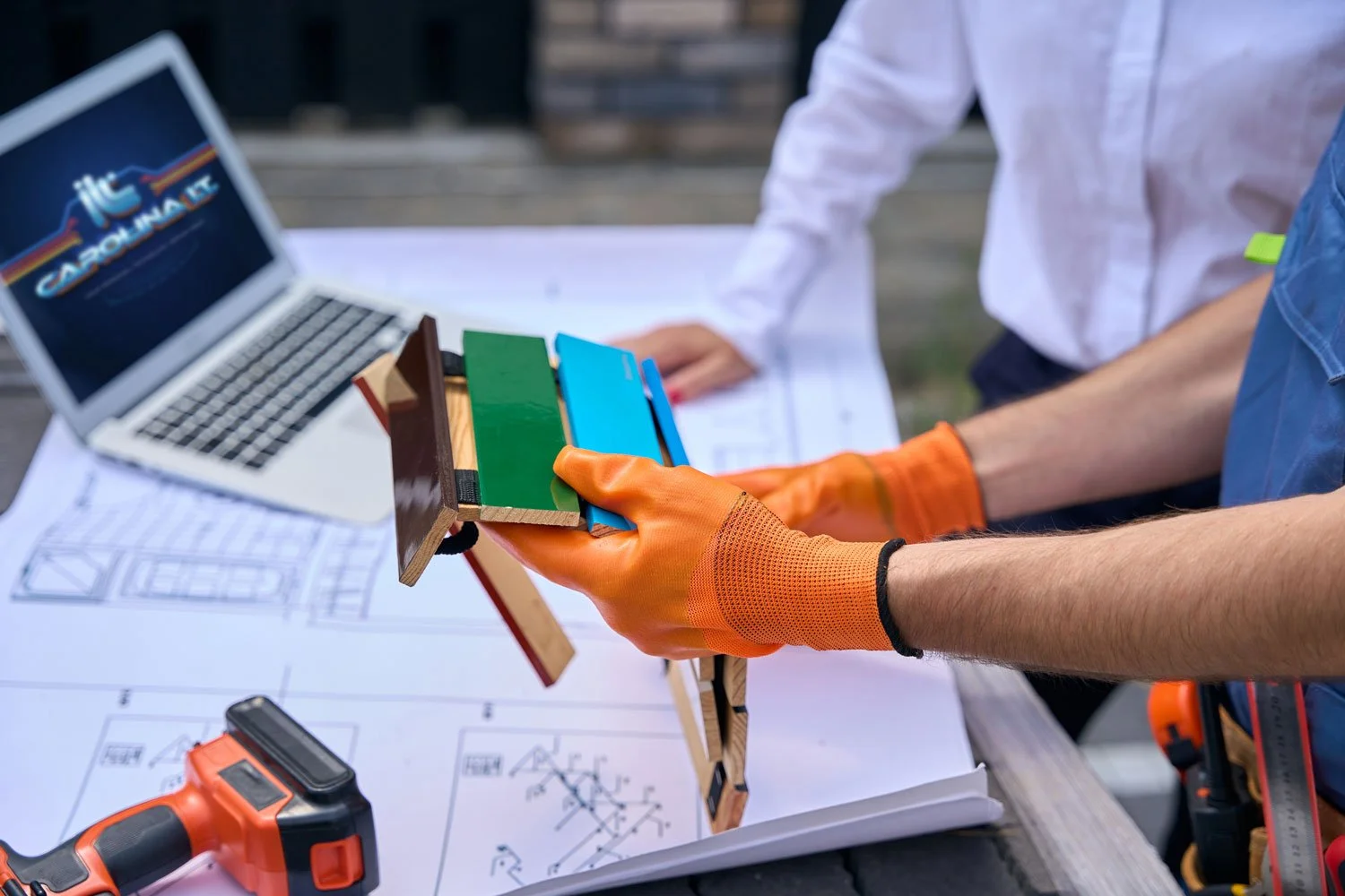 Two people working on a construction project, one holding color sample swatches, with a laptop, blueprints, and a power drill on the table.