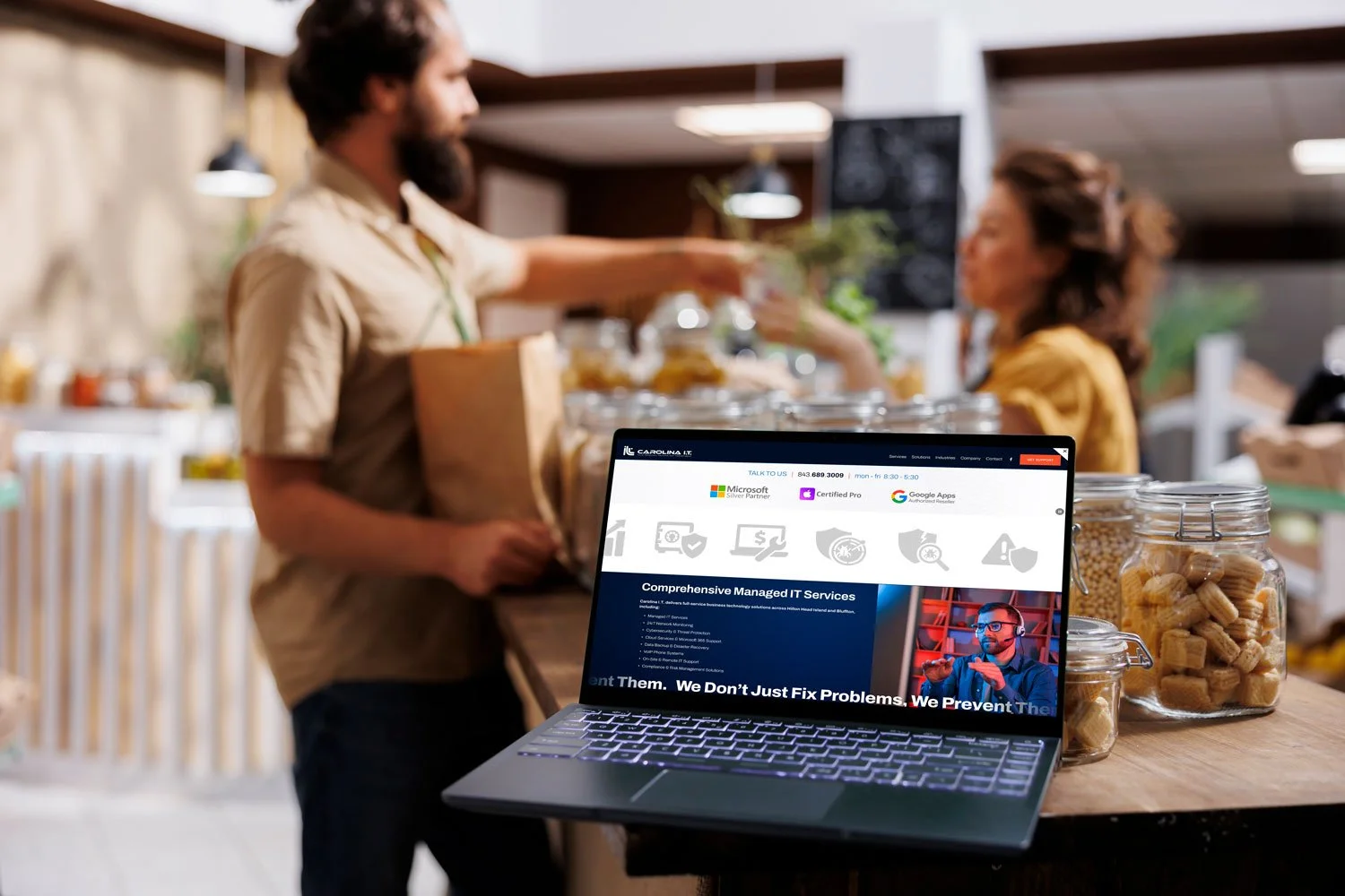 A coffee shop with a man handing a bag to a woman at the counter. In the foreground, a laptop displaying a managed IT services website is on the counter surrounded by jars of cookies and snacks.