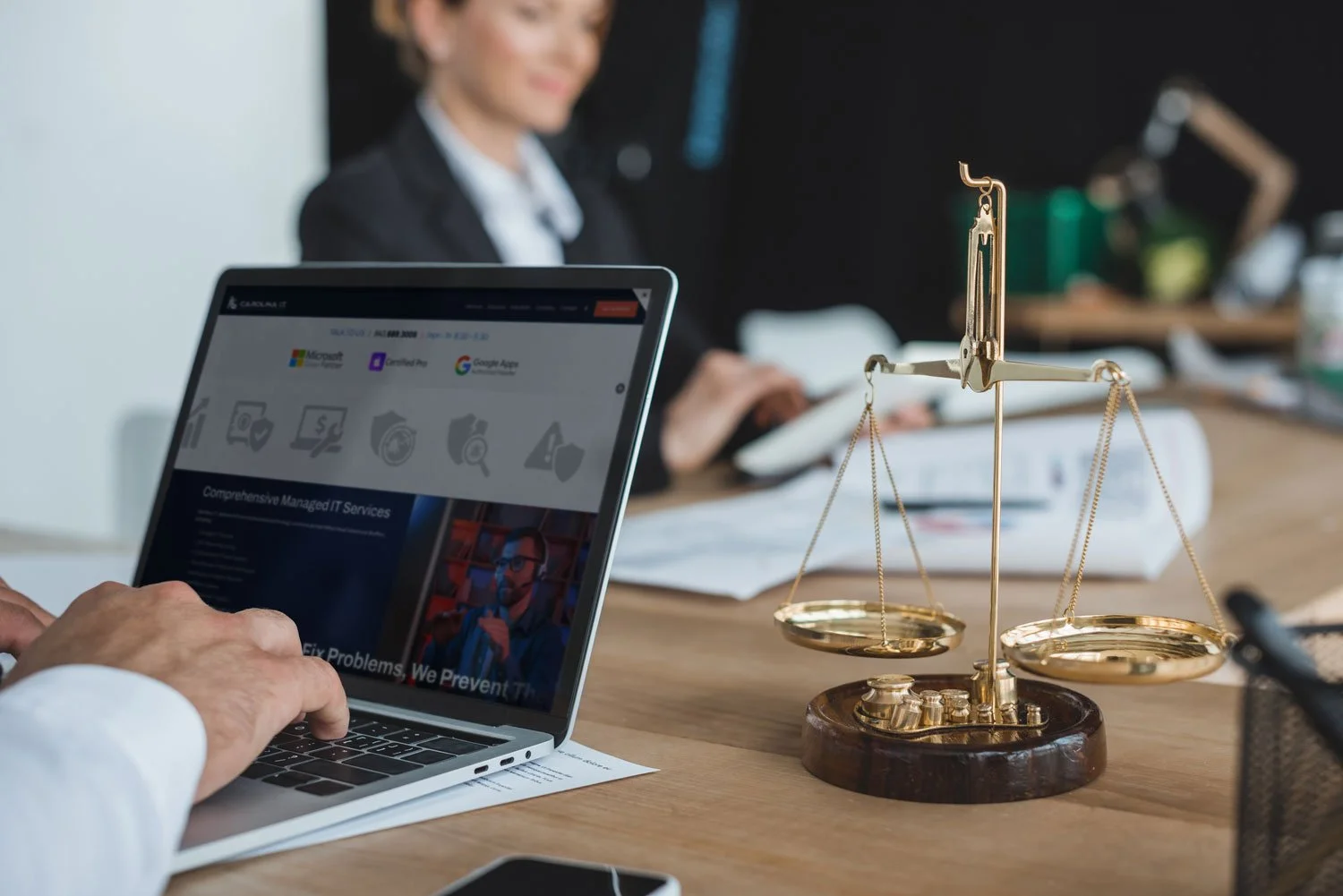 A person using a laptop on a wooden desk, with a golden scale of justice in the foreground and a woman in business attire blurred in the background.