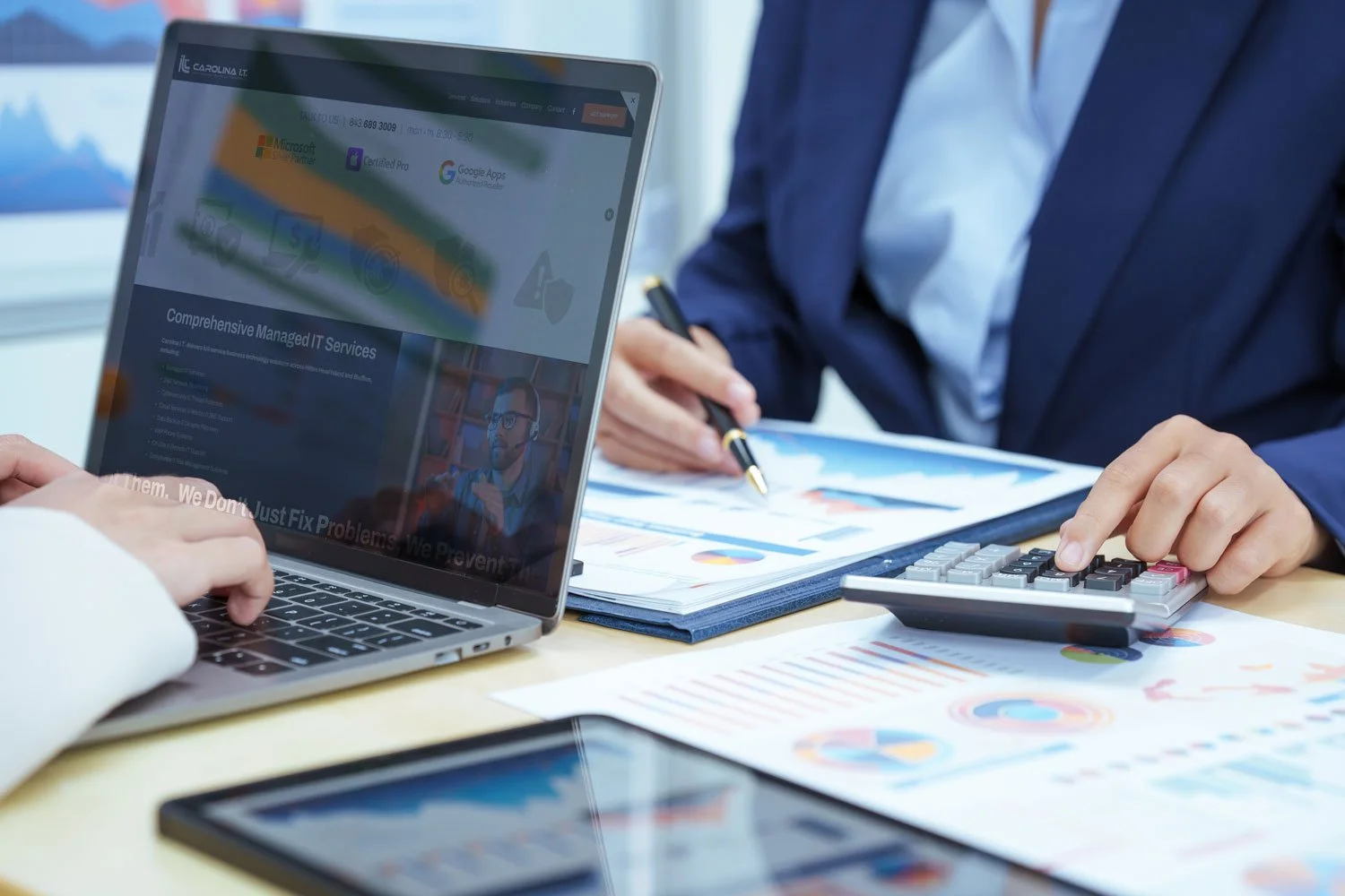 A person in business attire working on a laptop and another person using a calculator on a desk covered with financial charts and documents in an office setting.