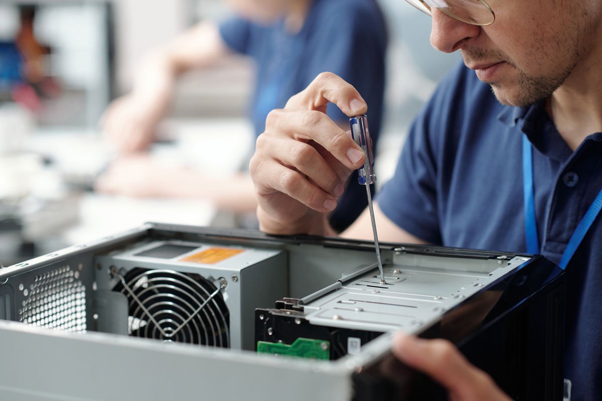 A technician repairing a desktop computer with a screwdriver in a workshop or office setting.