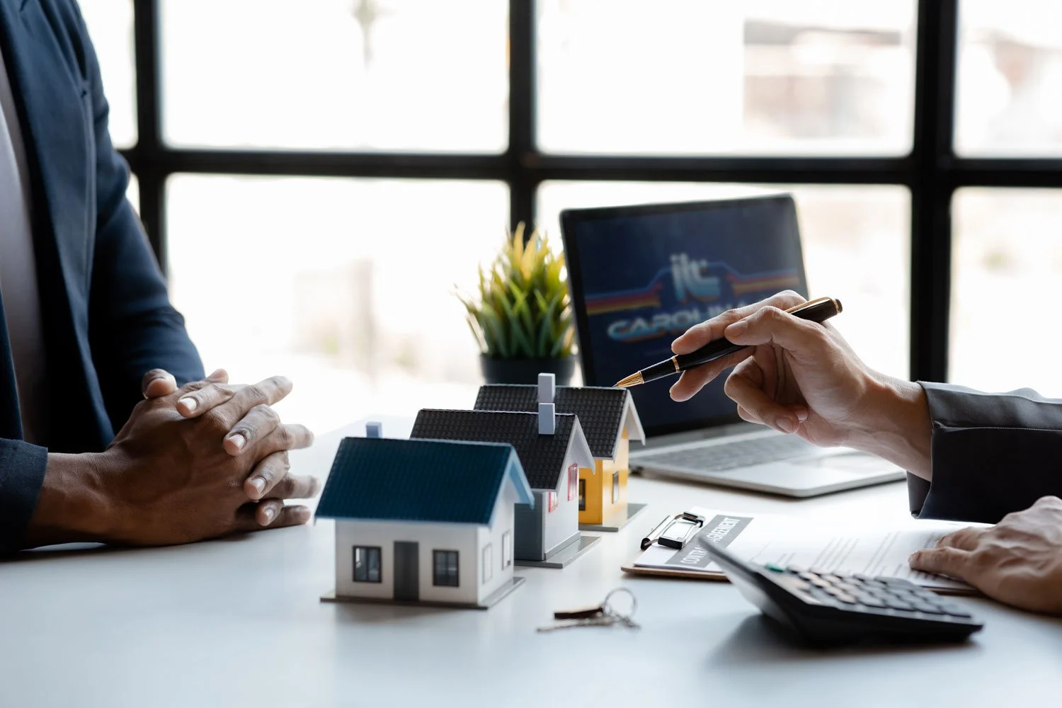 Two people are sitting at a table discussing real estate. One person has hands clasped on the table, while the other is pointing a pen at miniature house models. A laptop displaying a logo and a calculator are also on the table, with a clipboard and keys nearby. A small potted plant is in the background near a window.