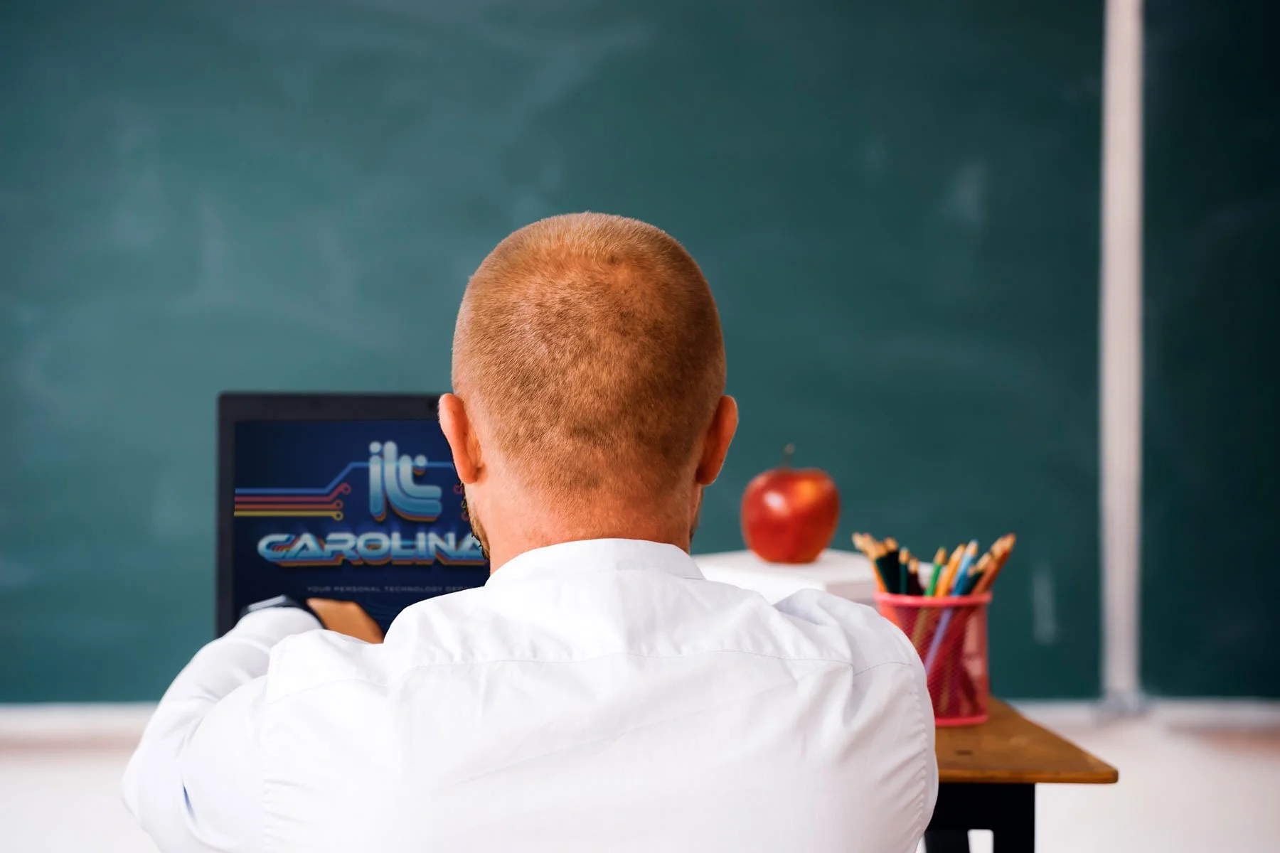 A person with a shaved head wearing a white shirt, sitting at a desk in front of a computer monitor with a colorful logo that reads 'Carolina.' There is a red apple, a pink container with pencils, and a chalkboard in the background.