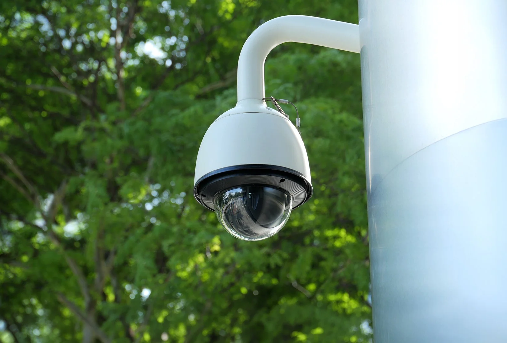 Security camera mounted on a pole outdoors with green trees in the background.