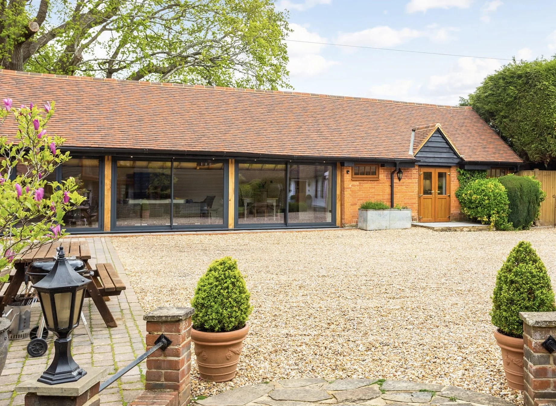 A brick house with a red tile roof and large glass sliding doors, surrounded by a gravel yard with potted plants and outdoor furniture.