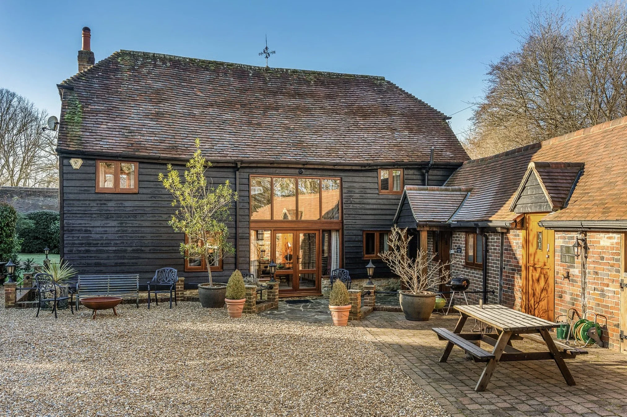 Photo of a backyard patio with a gravel surface, various potted plants, outdoor furniture, and a black and brick house with large windows and a steep roof.