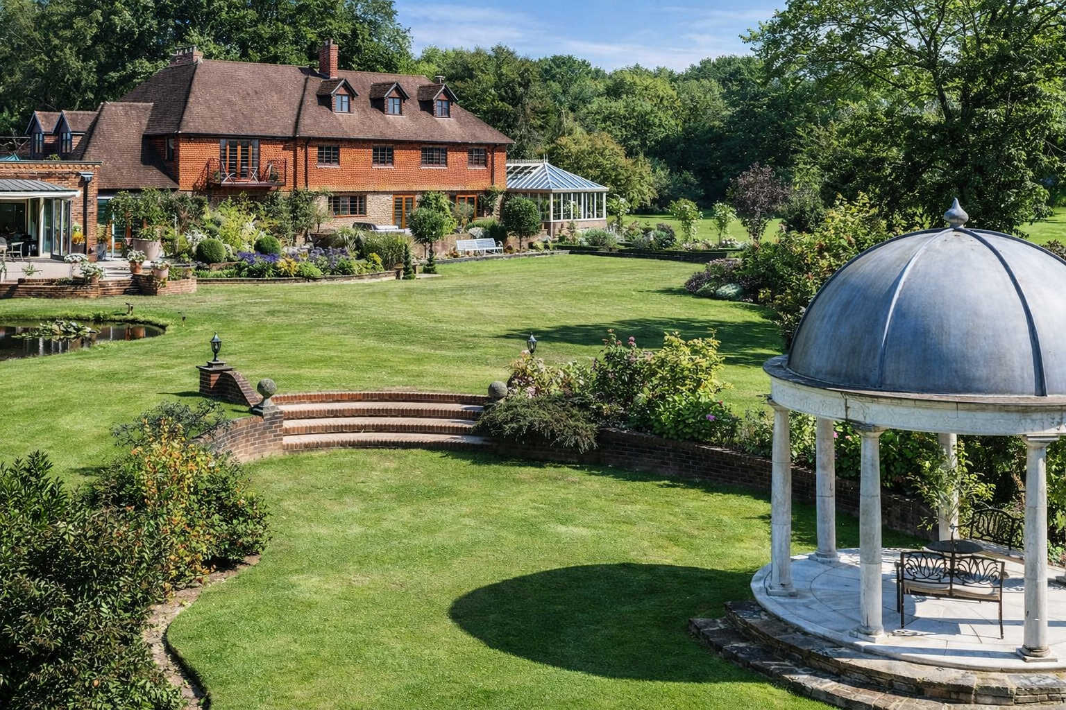 Large garden with well-maintained grass, a pond, and decorative landscaping. A mansion-style house with multiple stories, dormer windows, and a greenhouse is in the background. A white, round gazebo with columns and benches is in the foreground.
