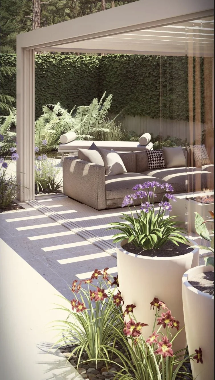 A cozy outdoor patio area with a gray sofa and various cushions, surrounded by lush green plants and flowers in pots, with sunlight casting shadows on the tiled floor.