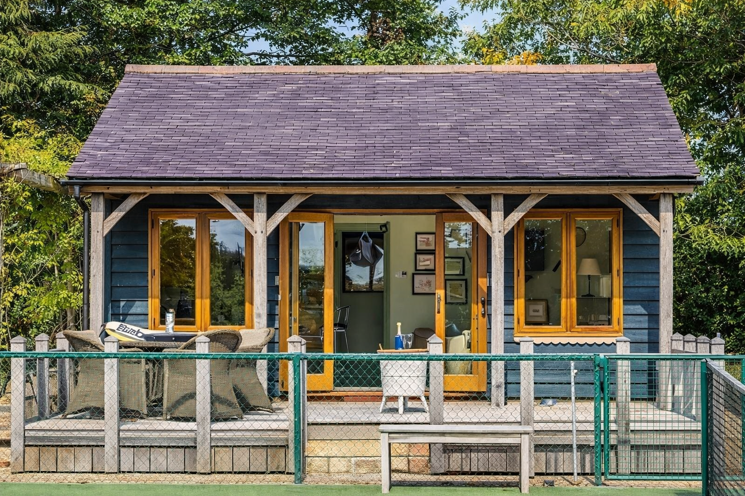 Small house with a dark blue exterior, yellow window and door frames, and a gray shingle roof. It has a front porch with outdoor furniture and is surrounded by green trees and a fence.