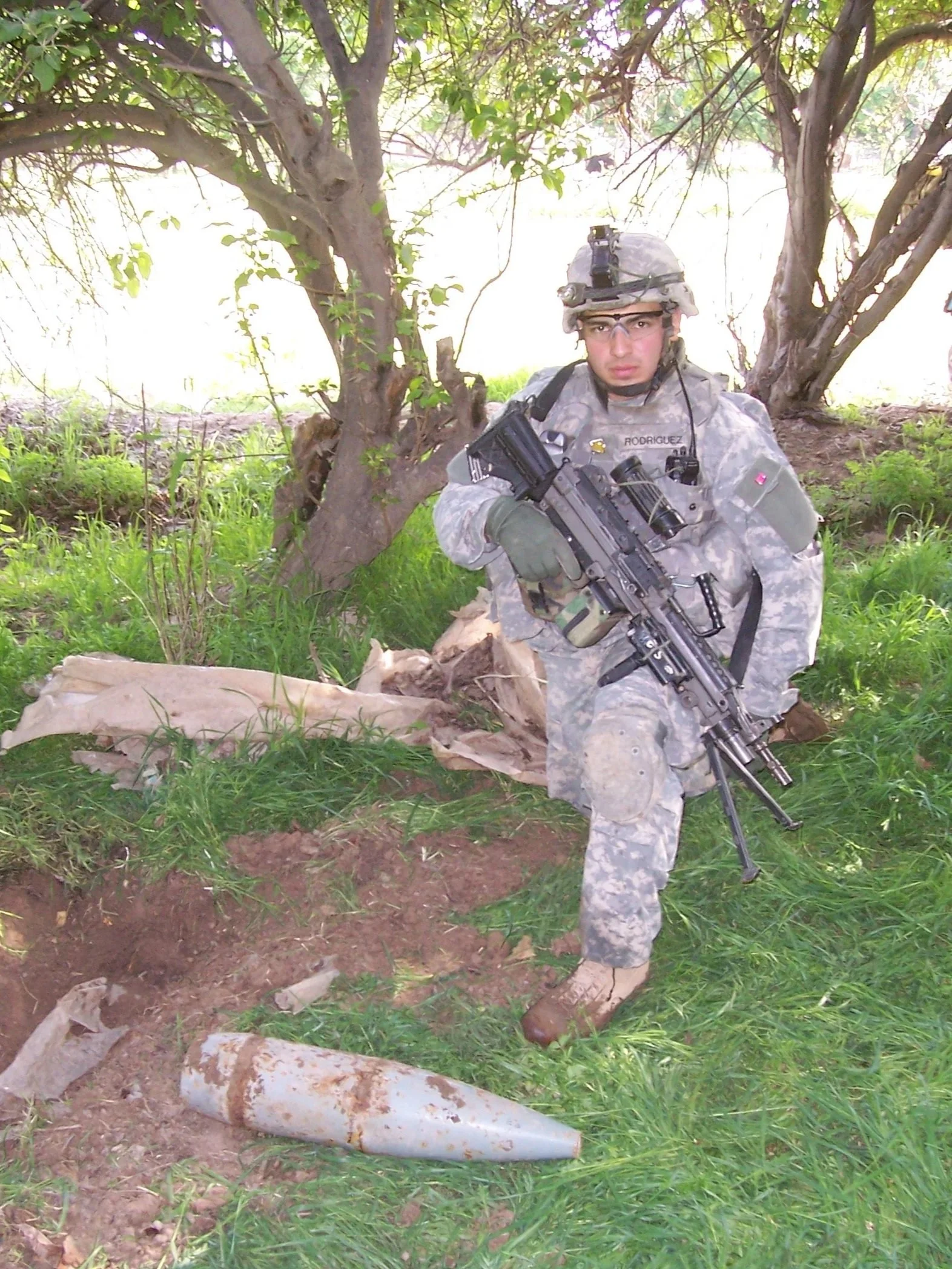 A soldier in camouflage uniform kneeling on grass next to a tree trunk, holding a rifle, with a large artillery shell lying on the ground nearby.