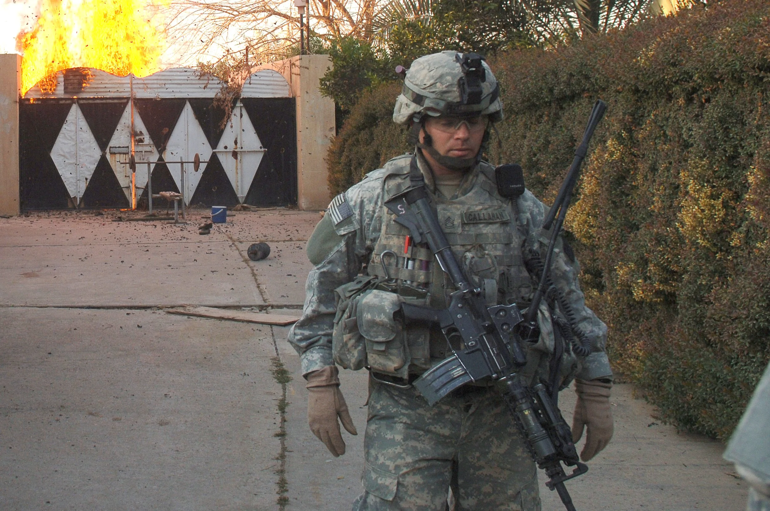 A soldier in tactical gear standing outdoors, holding a rifle, with a background of a burning structure and debris scattered on the ground.