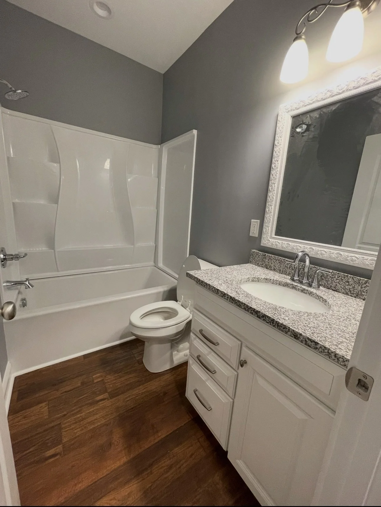 Bathroom with vanity, granite countertop, oval mirror, toilet, bathtub with shower, and wood flooring.