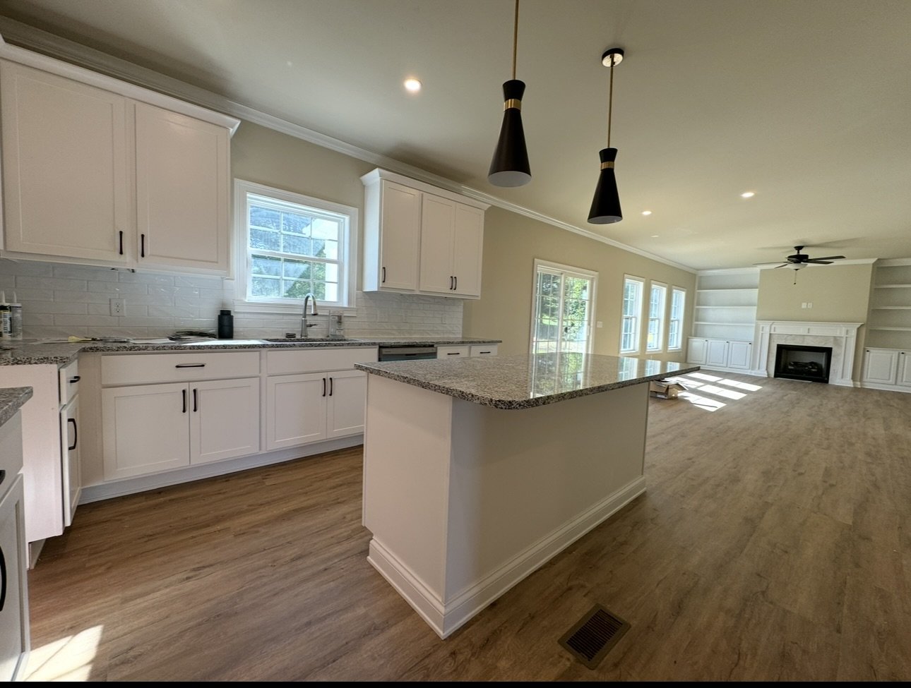 A spacious open-concept living area with a kitchen featuring white cabinets, granite countertops, and a central island, overlooking a living room with a fireplace and built-in shelves, illuminated by multiple windows and ceiling lights.