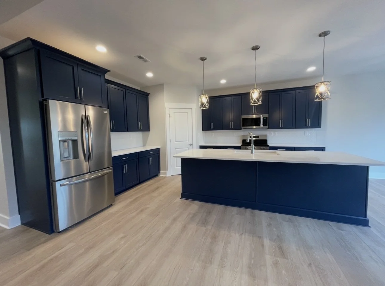 Modern kitchen with navy cabinets, stainless steel refrigerator, island with white countertop, pendant and recessed lighting, hardwood floors.