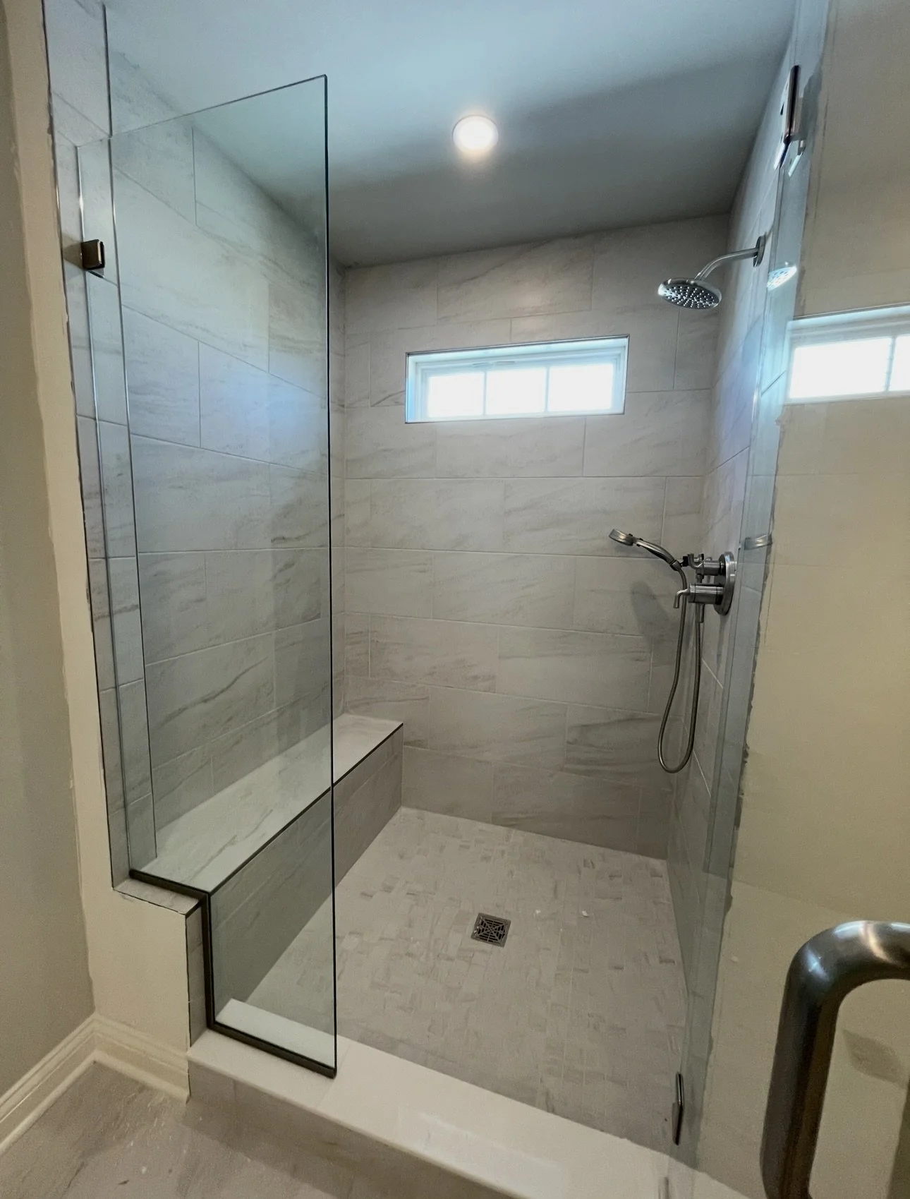 Modern walk-in shower with glass enclosure, beige tile walls, a small horizontal window, rainfall showerhead, hand-held shower sprayer, and a built-in bench inside the shower.