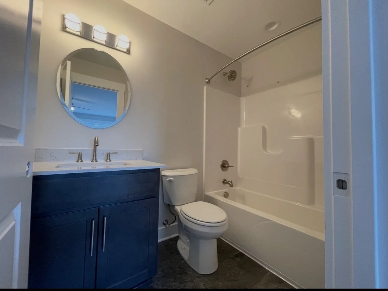 A small bathroom with a white bathtub and shower combination, a toilet, a blue vanity with a sink, a round mirror, and a light fixture above the mirror.