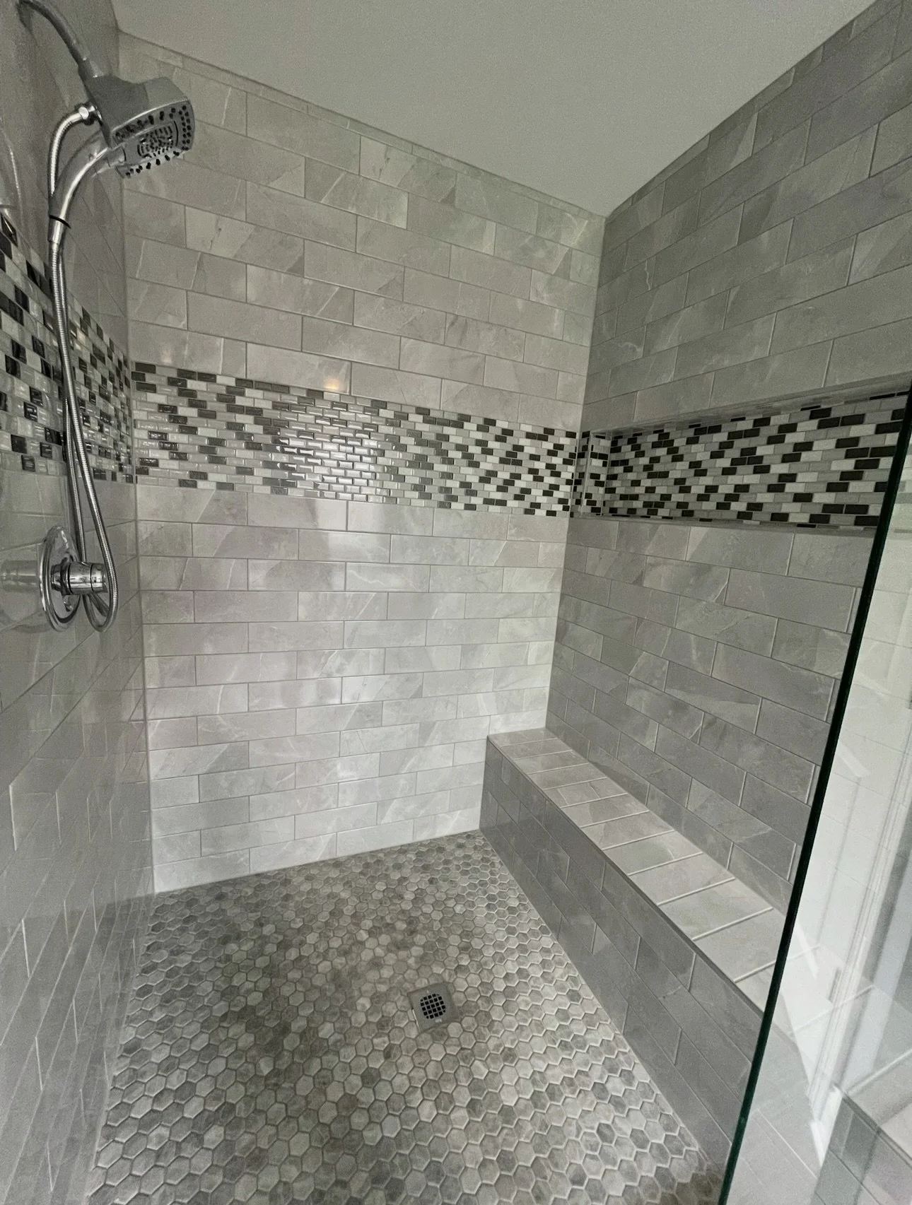 A modern walk-in shower with gray subway tiles, a decorative strip of small black, white, and gray mosaic tiles, a built-in bench, and a ceiling-mounted shower head.