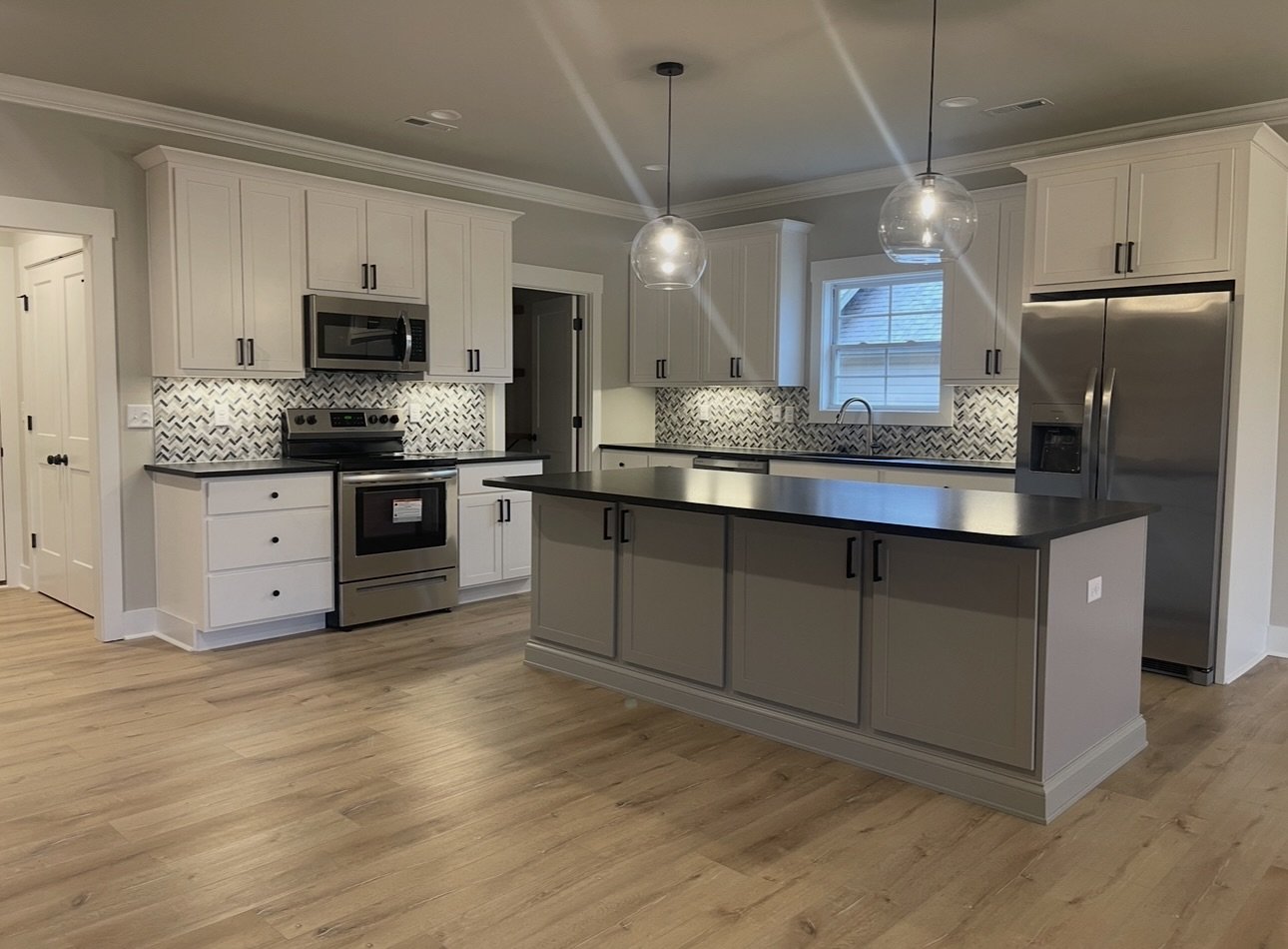 Modern kitchen with white cabinets, black countertops, stainless steel appliances, and a patterned backsplash. An island with dark gray cabinets is in the center, and light wood flooring is throughout.