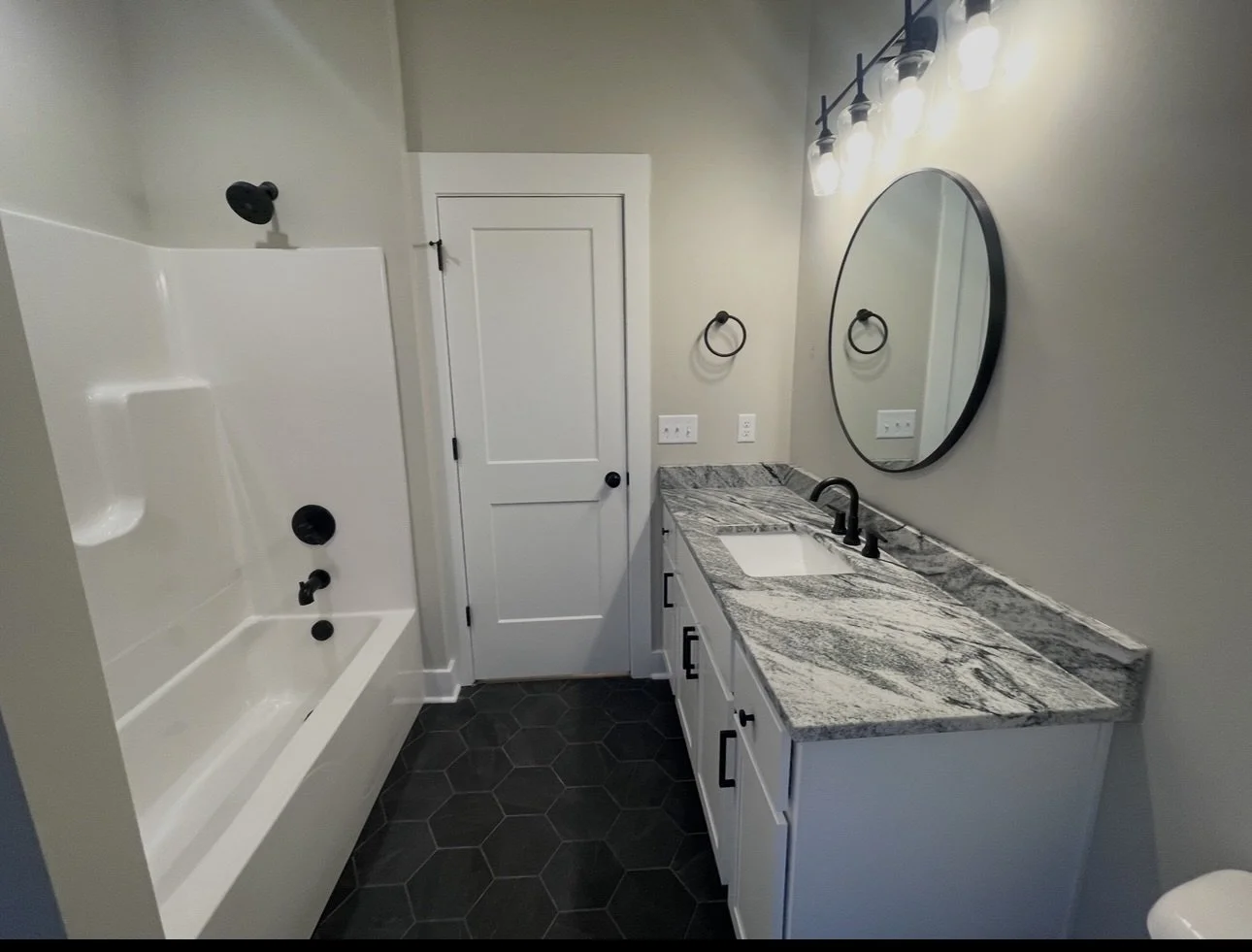 Modern bathroom with a white bathtub and shower, black fixtures, a marble countertop vanity with black hardware, an oval mirror, and black hexagonal floor tiles.